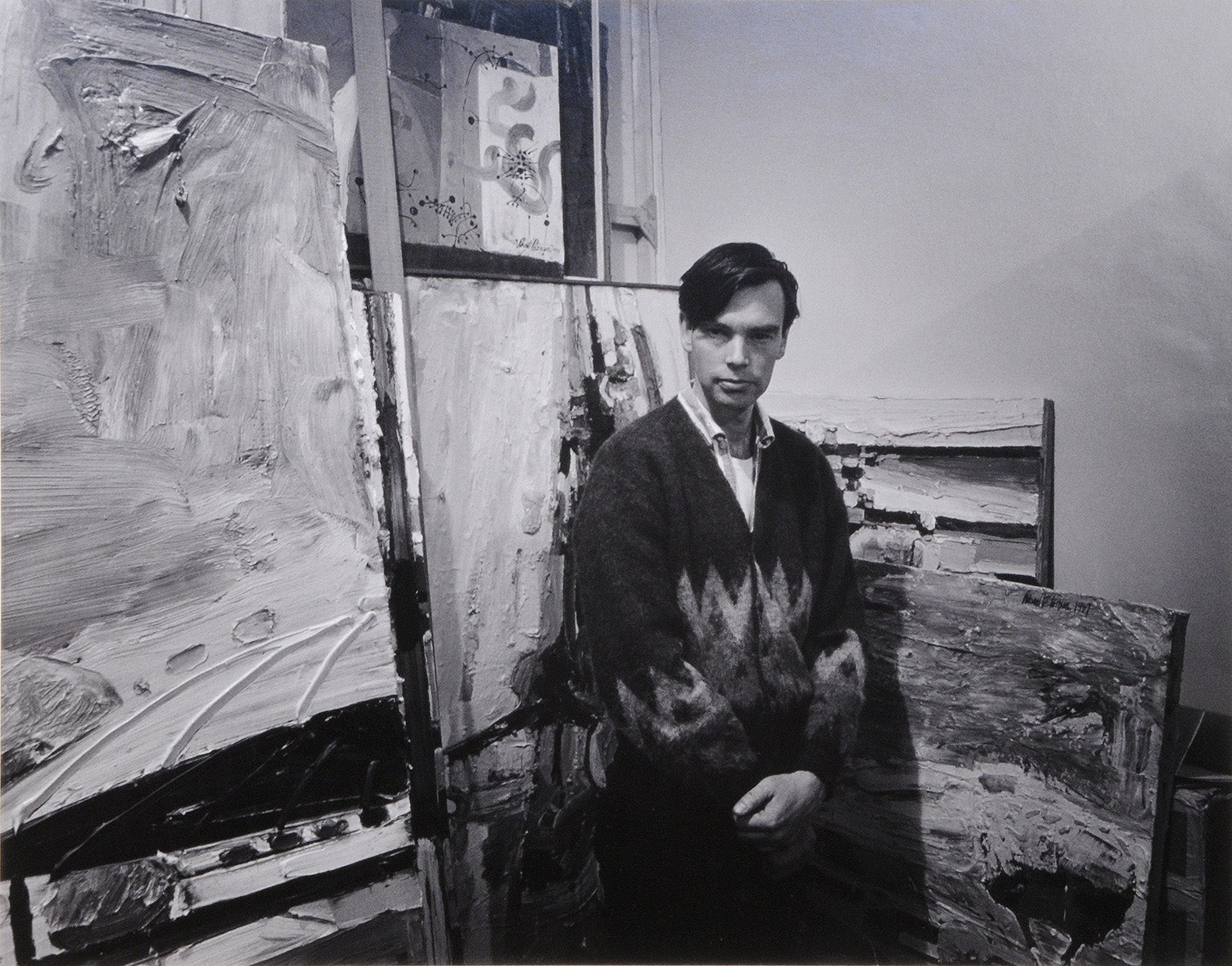 A man in a sweater stands in an artist's studio surrounded by abstract paintings.