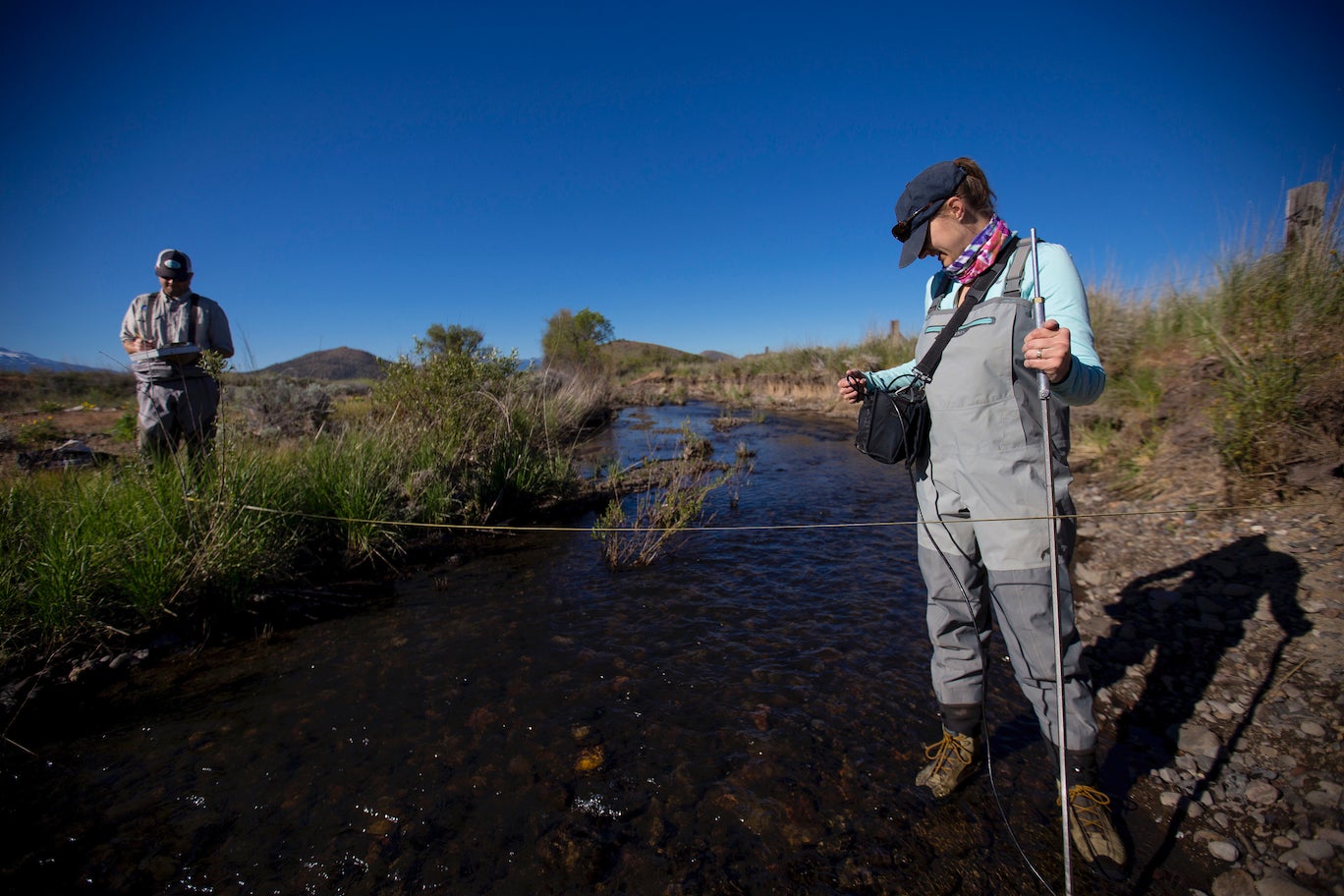 Woman scientist stands in stream with monitoring equipment with male scientist in background