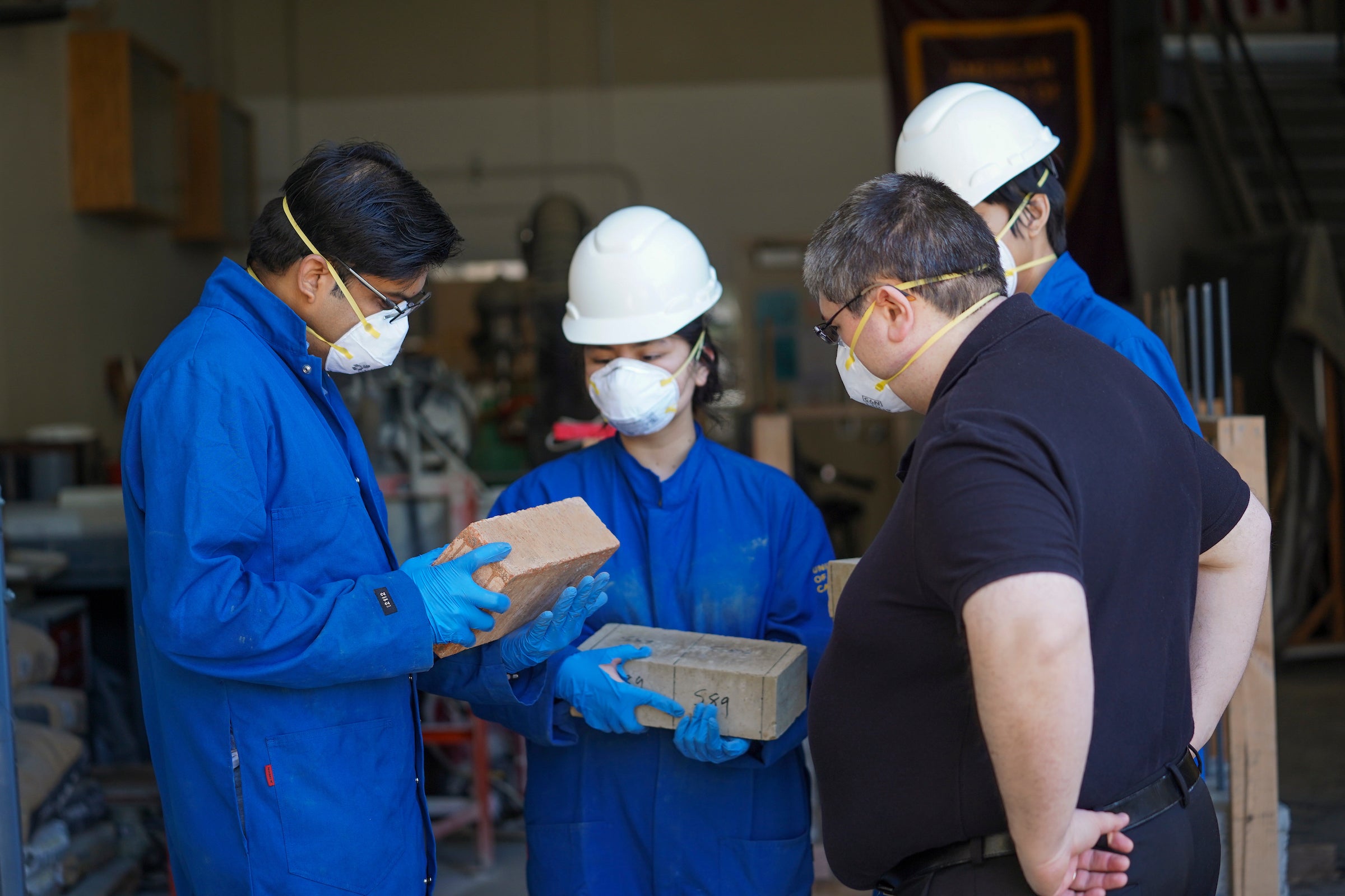 Four scientists in blue lab coats huddle together looking at compressed earth block they made. 