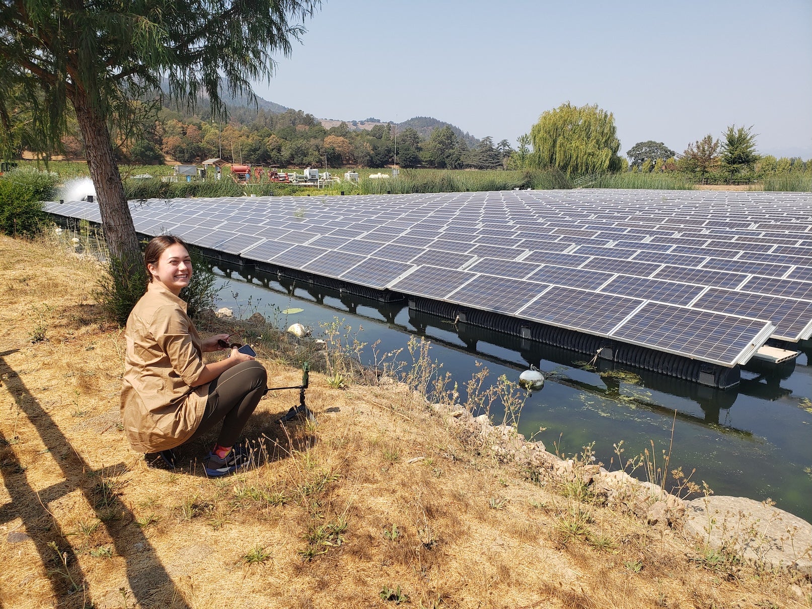 Emma Forester in tan shirt and pants crouches on gold-colored grasses alongside a pond with a floating solar photovoltaic system and smiles at the camera.