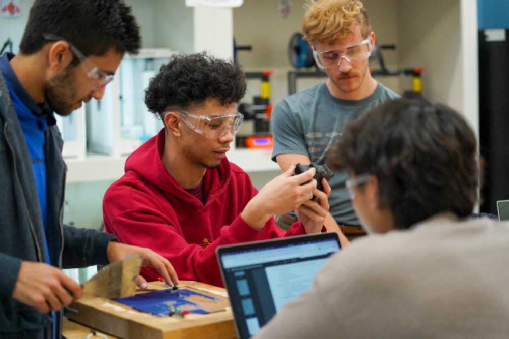 A group of four students in the lab wearing goggles discusses a project. One student in the center holds and examines a small, specific component.