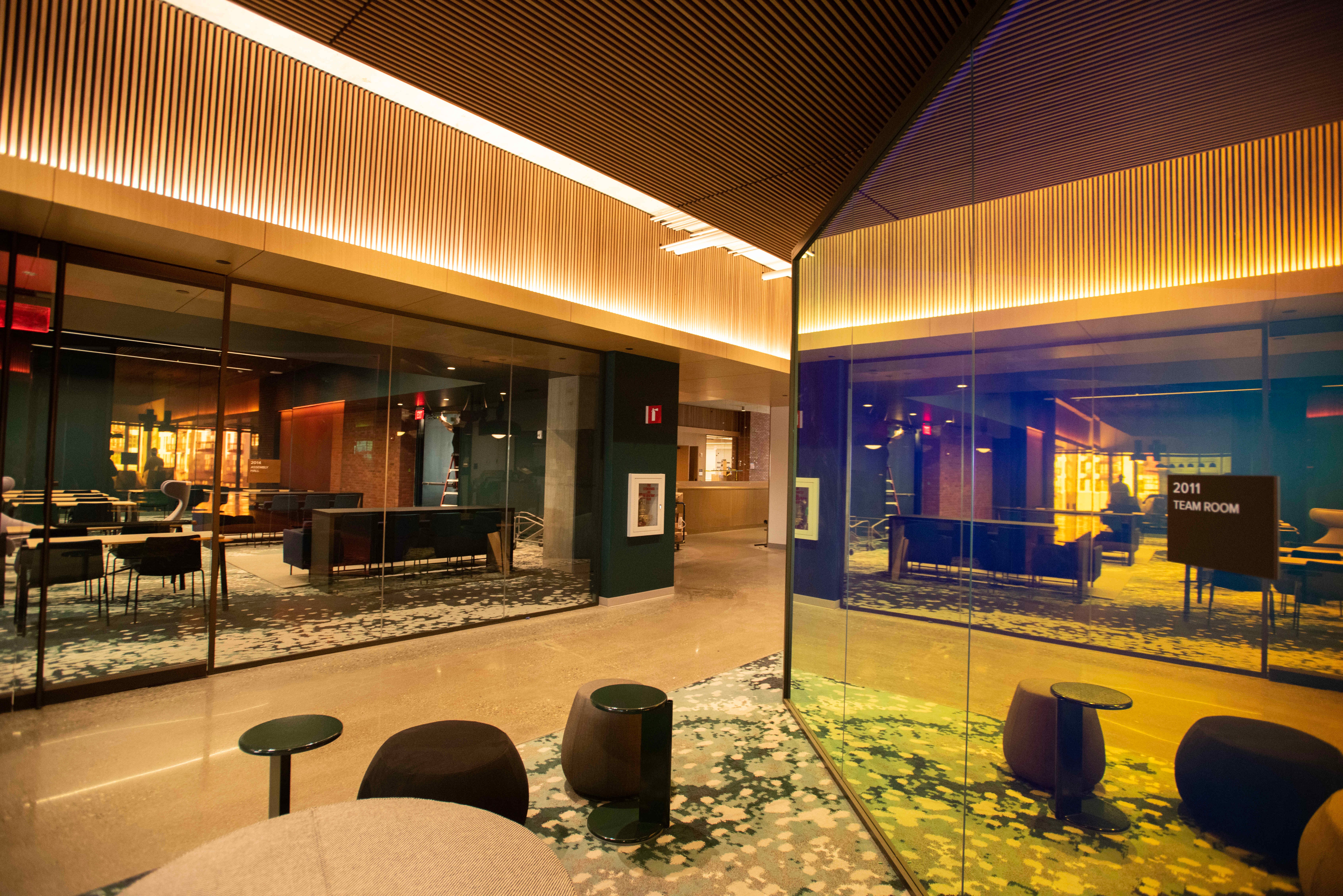 An angular view of two brightly lit, glass offices converging in a carpeted common space with chairs and a table.