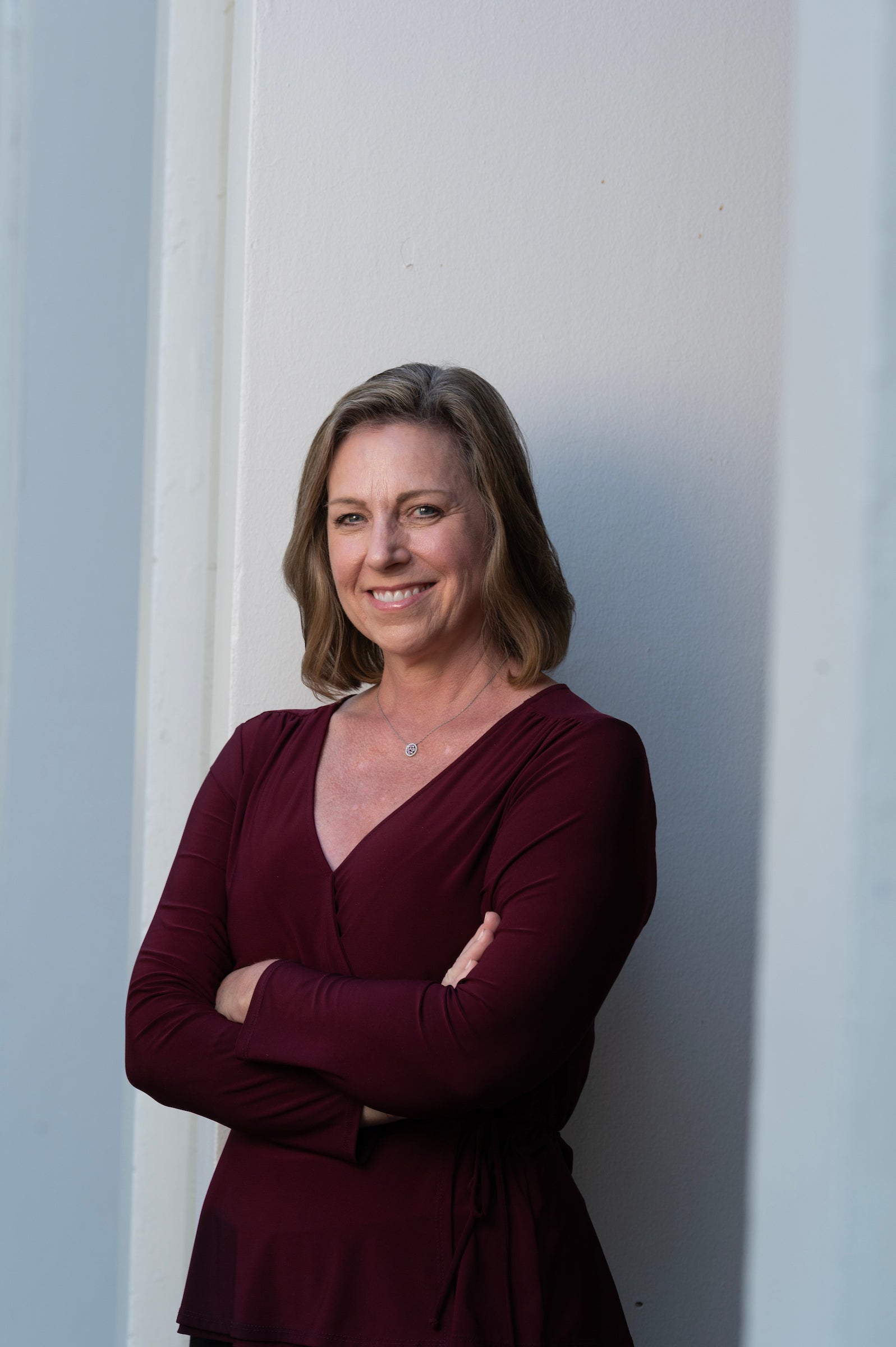 outdoors against a white building, a woman stands with their arms crossed, smiling at the camera. 