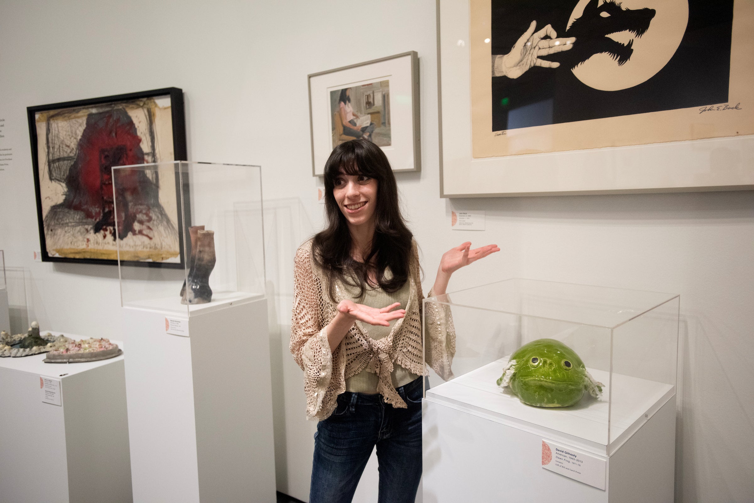 A woman stands in an art gallery, gesturing near a green sculpture and various framed artworks.
