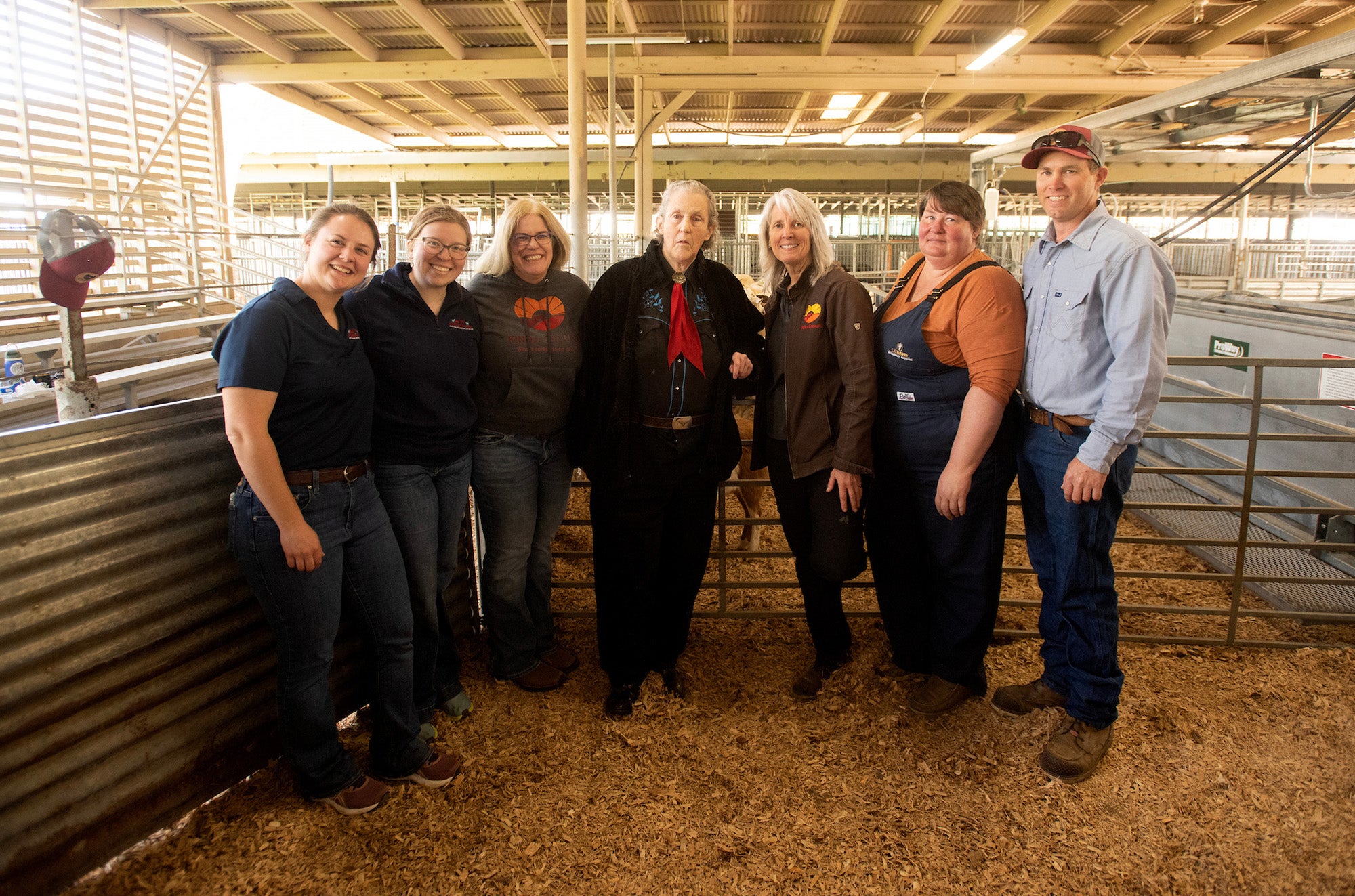 Seven people pose together inside a barn, smiling in front of animal pens. Left to right: Ashlynn Kirk, Karly Anderson, Cassandra Tucker, Temple Grandin, Jen Walker, Fauna Smith, Nathan Medlar. (Gregory Urquiaga / UC Davis)