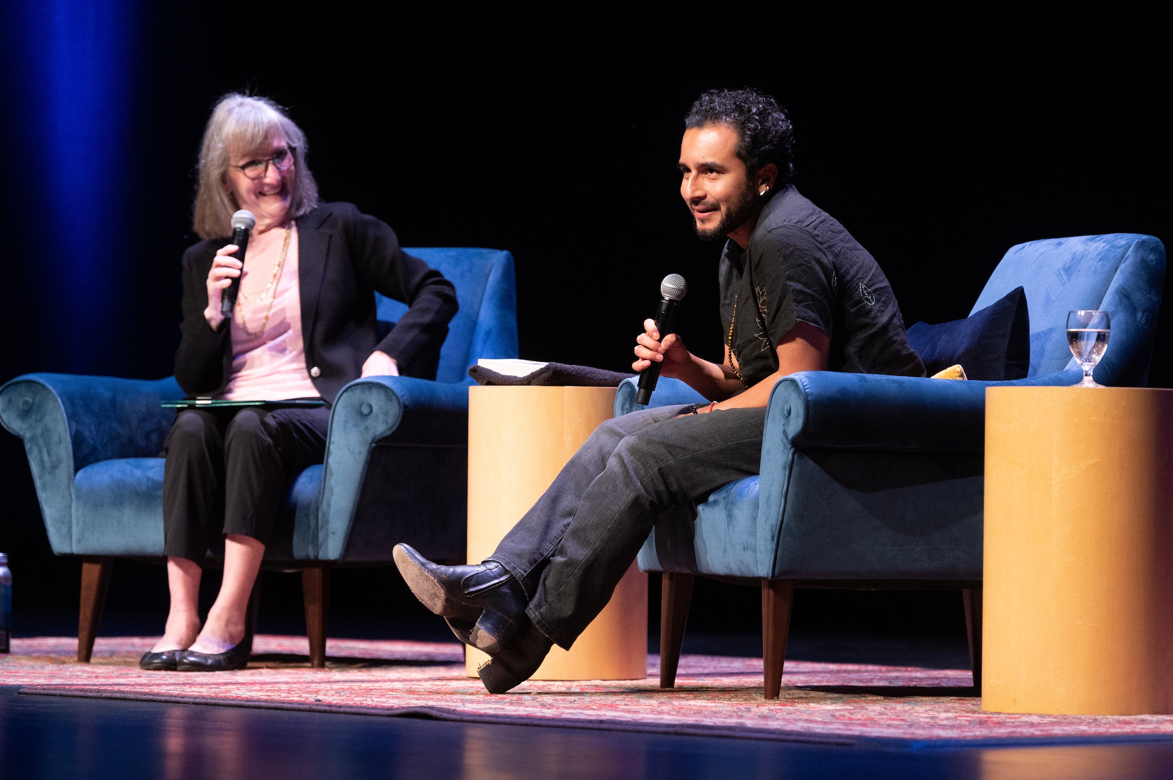 Two presenters -- Donna Apidone to the left and Javier Zamora to the right -- sit on a stage in blue chairs, engaging during a discussion.