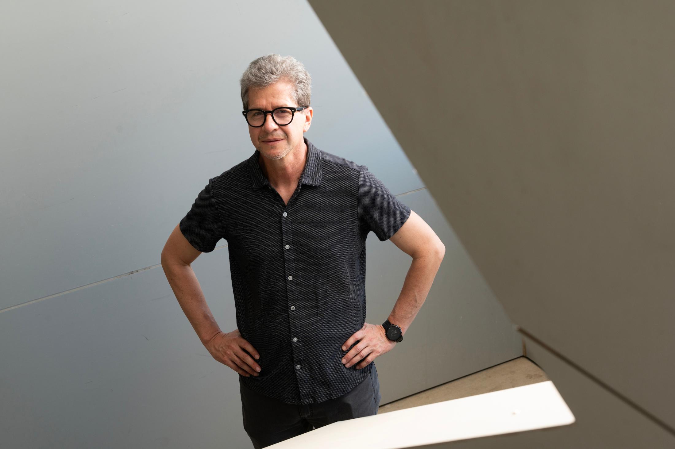Middle-aged man with gray hair and glasses in black shirt, hands on hips in angular modern space