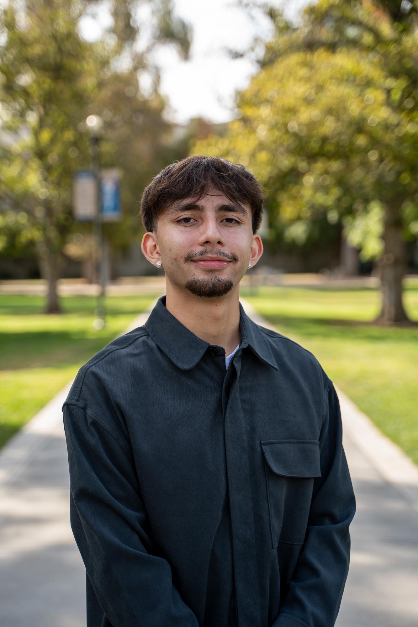 A student smiles and stands on a sunlit path near the UC Davis Quad. Trees with green leaves and a blurred campus sign are visible in the background.
