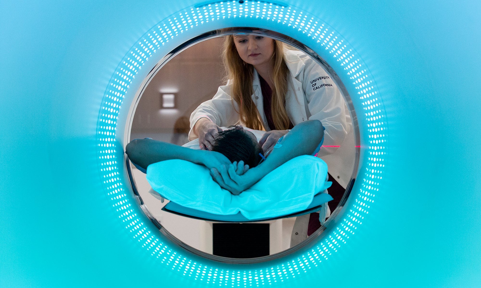 In a circular opening, a long-haired woman in a white lab coat bends over a person lying down with the top of their head towards the camera. Blue color fills the rest of the field outside the circle. 