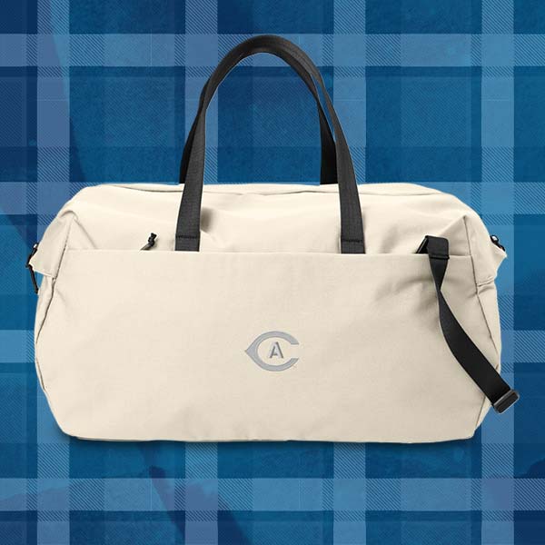 a cream-colored duffel bag with hand and shoulder straps in photographed in front of an Aggie Blue backdrop. The UC Davis Athletics logo is on the duffel bag. 