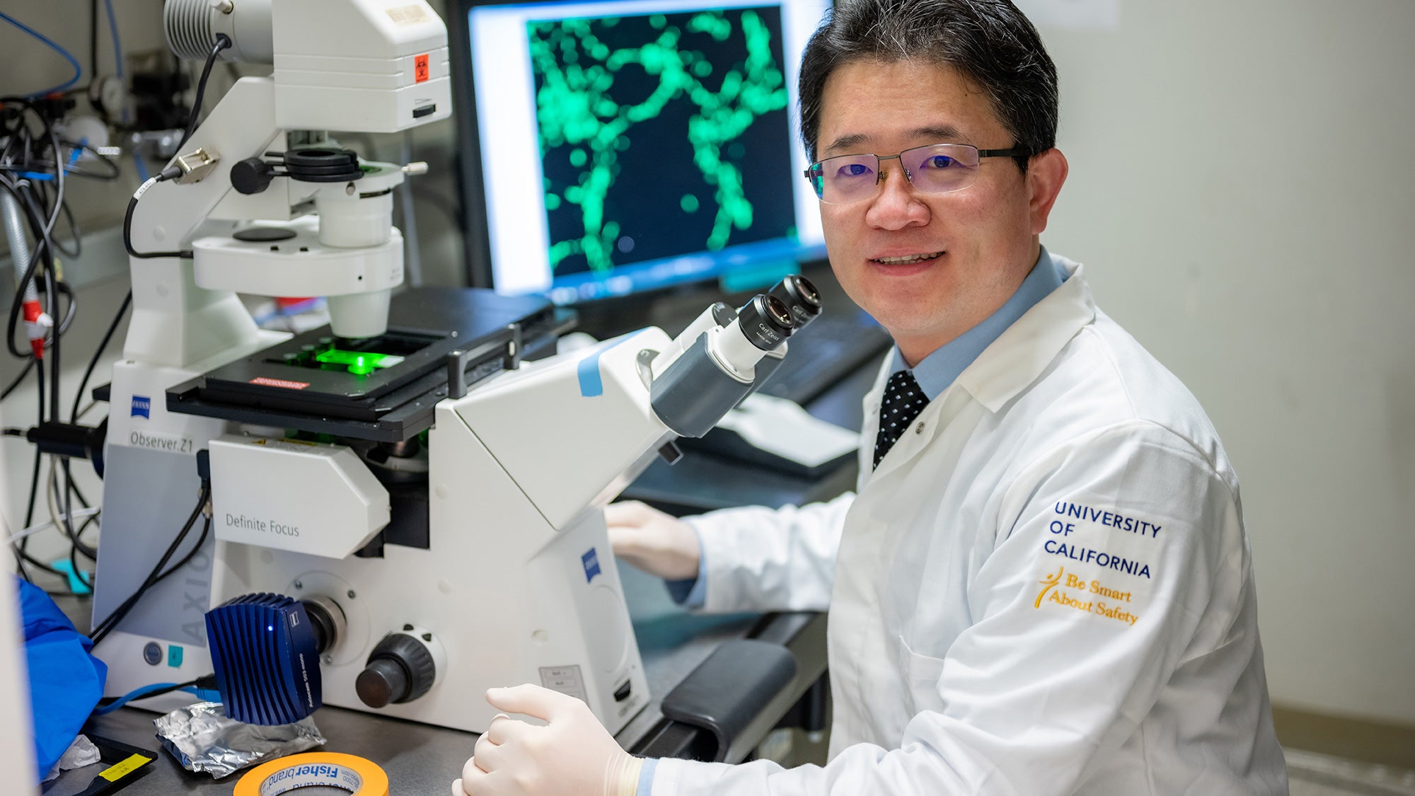 Researcher in lab coat sits with a microscope and computer screen