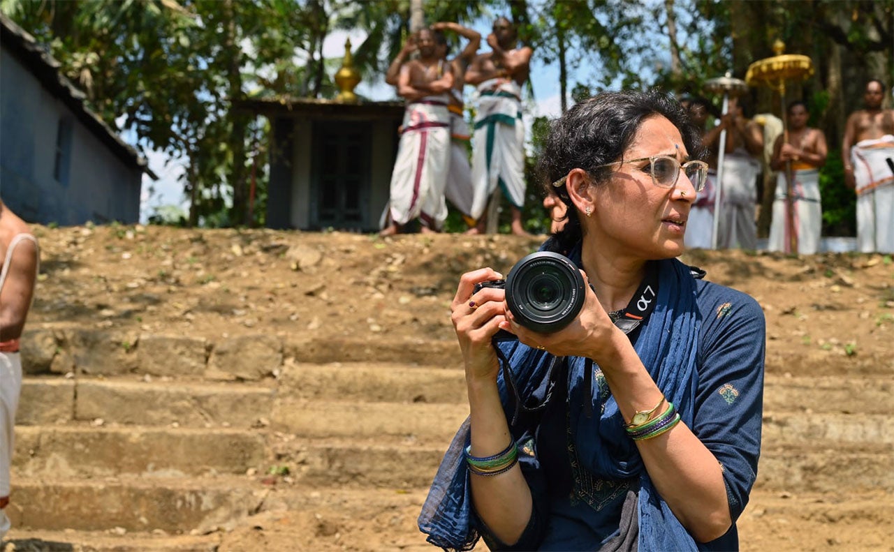 A woman in a blue outfit holds a camera while observing an event in the background.