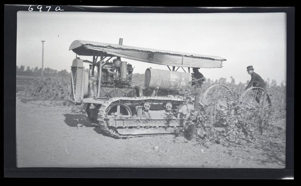 Two workers using a vintage tractor in a field, with crops in the foreground.