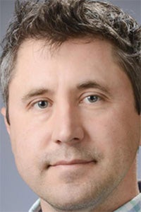 A close-up portrait of a man with short grayish hair and blue eyes, looking directly at the camera.
