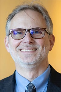 Smiling man with glasses and a suit, set against a warm, soft-focus background.