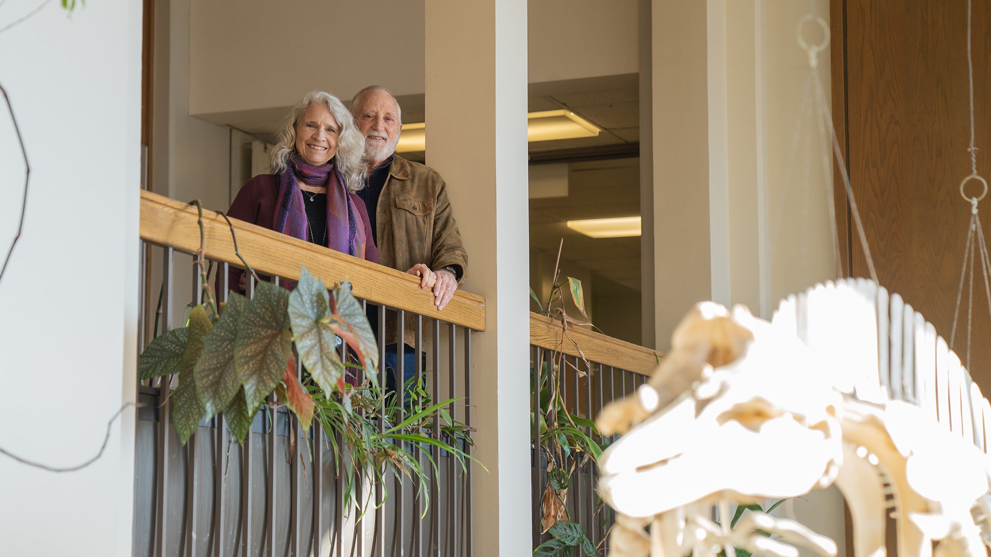 Two smiling individuals stand together on a balcony surrounded by indoor plants.