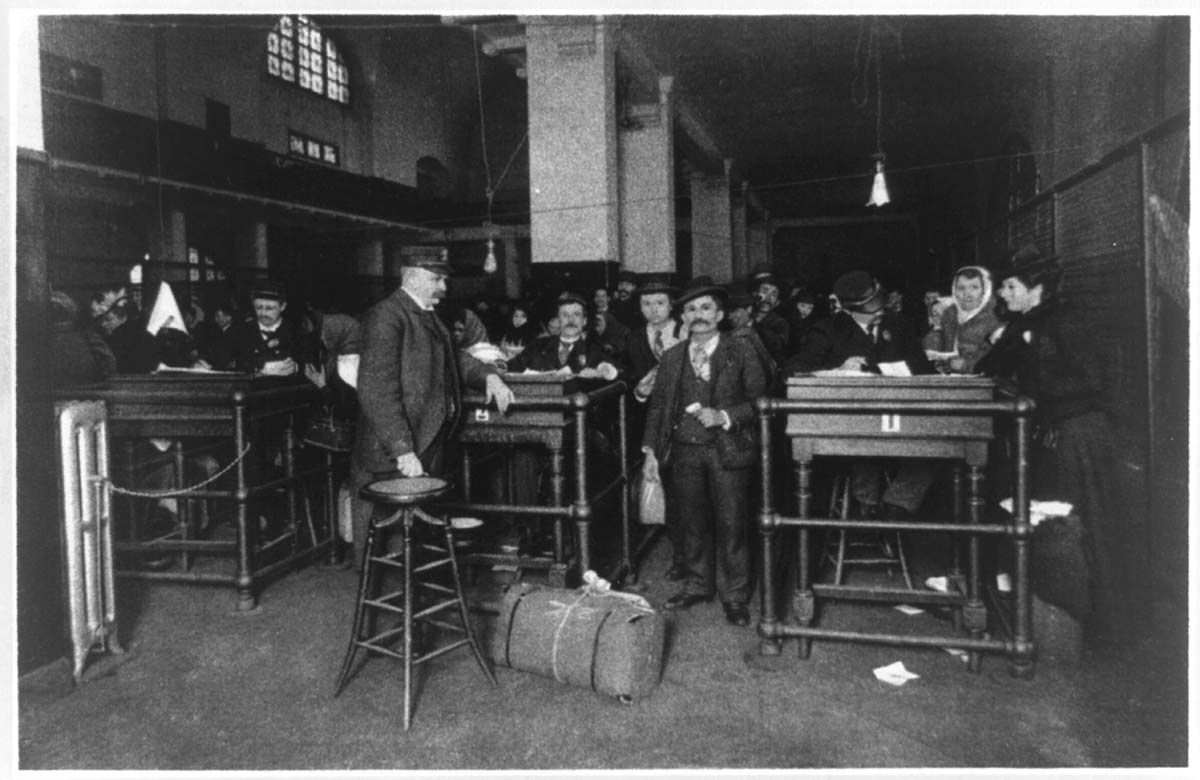 Historic black-and-white photograph of the final discharge area at Ellis Island in the early 20th century. Immigrants, many in period clothing and hats, stand in line or speak with officials at tall wooden desks. Some people hold documents, while luggage and parcels are visible on the floor. The room is dimly lit with bare lightbulbs hanging from the ceiling, and large arched windows are partially visible in the background.