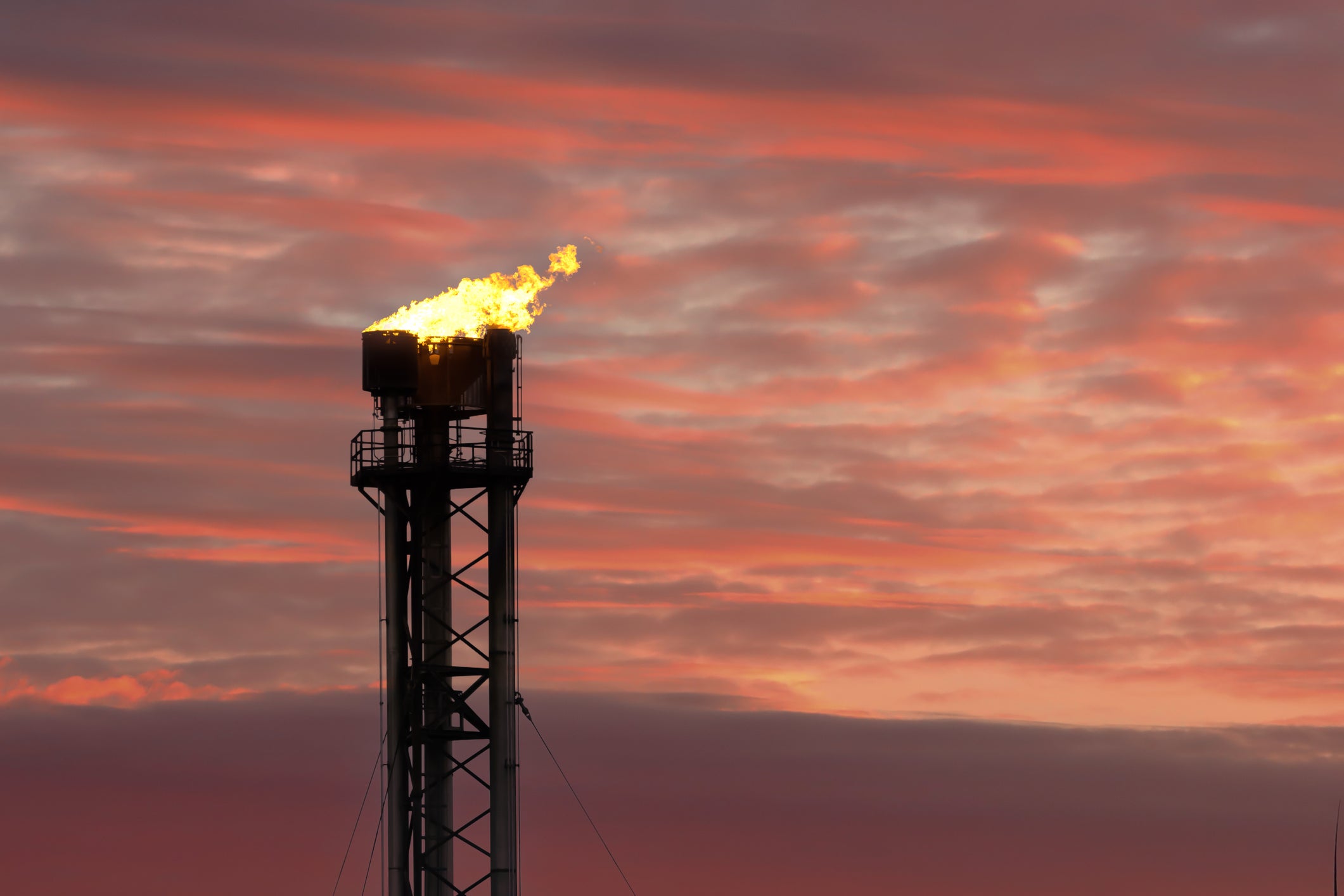 A firey gas flare atop a gas plant silouetted against a sunset sky.