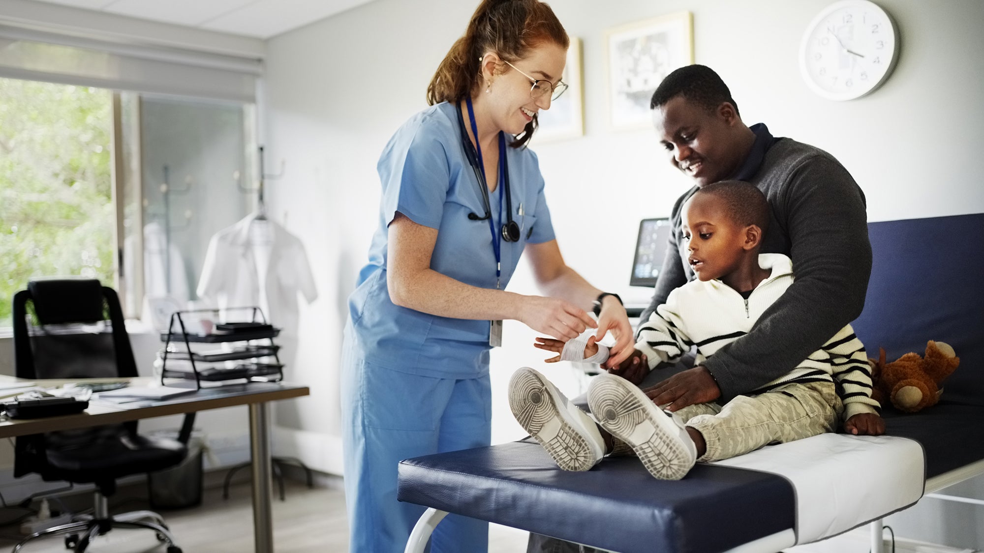 Doctor wrapping gauze on injured boy's hand with father comforting in clinic