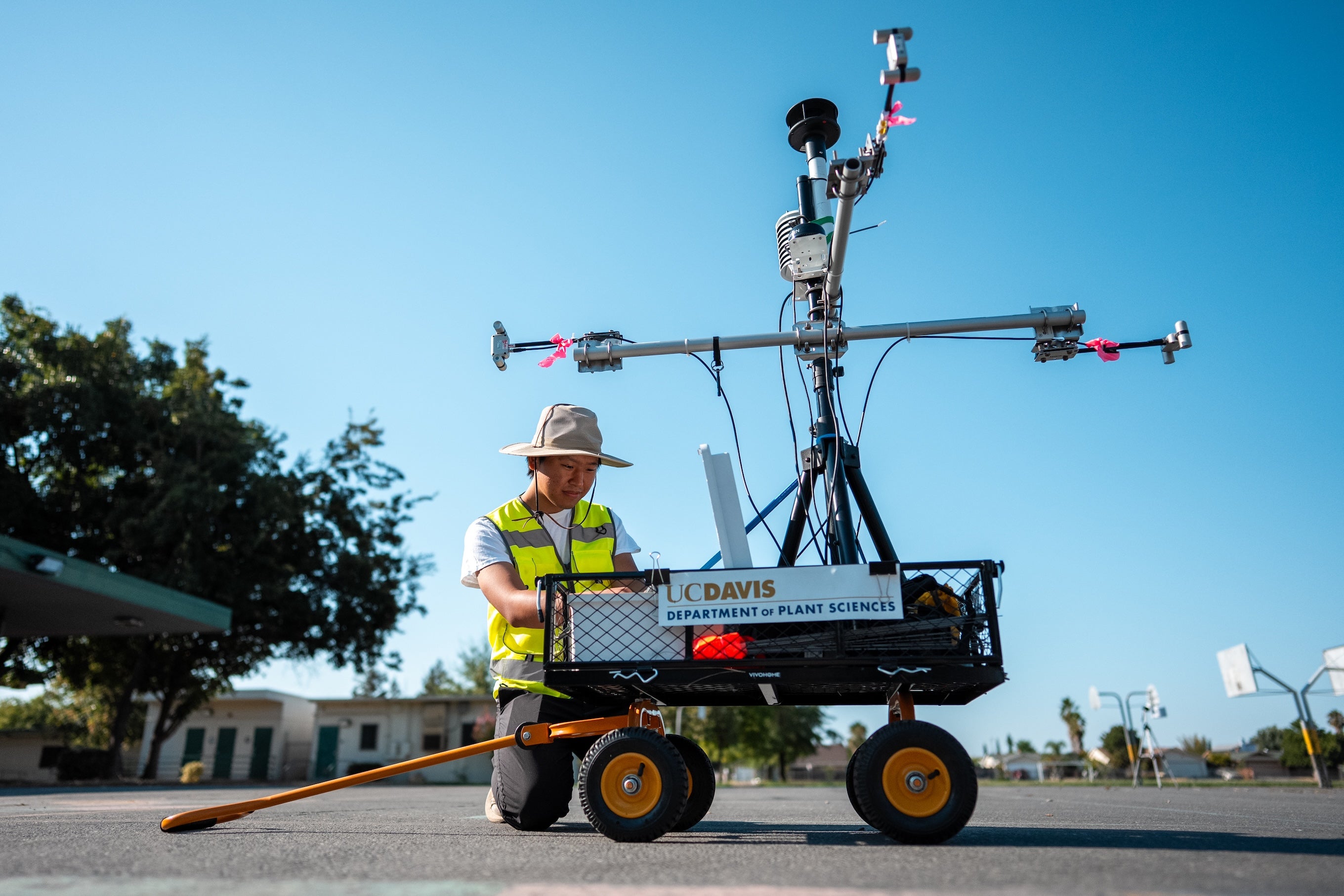 Tyler Reece Wakabayashi kneels next to a cart loaded with scientific instruments on blacktop under a blue sky with tree in background