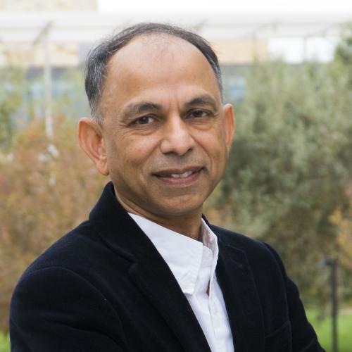 man smiling in a garden setting, wearing a black blazer over a white shirt.M