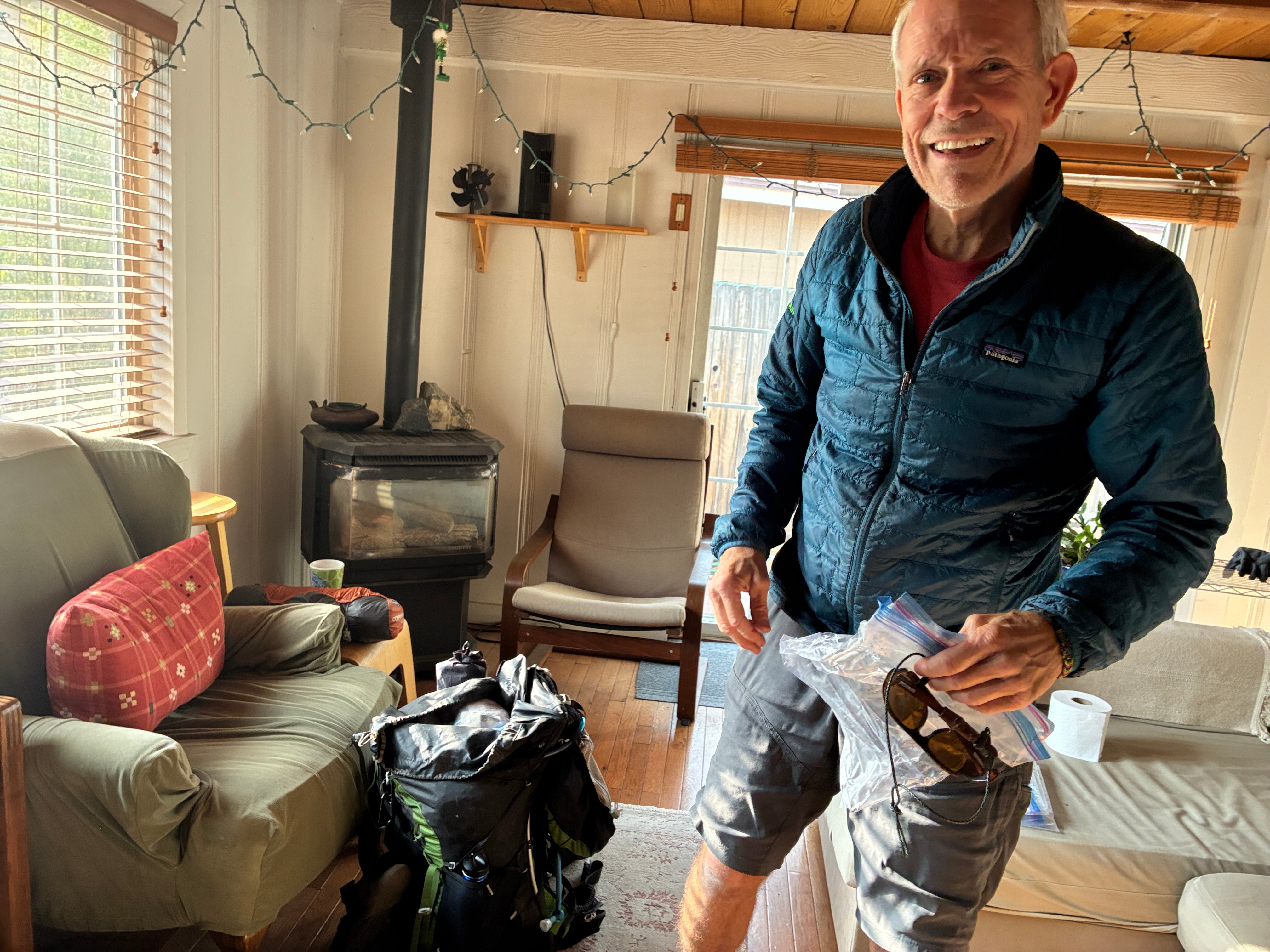 Man in blue puffer jacket stands smiling in a living room while packing for a trip with wood furnace in background