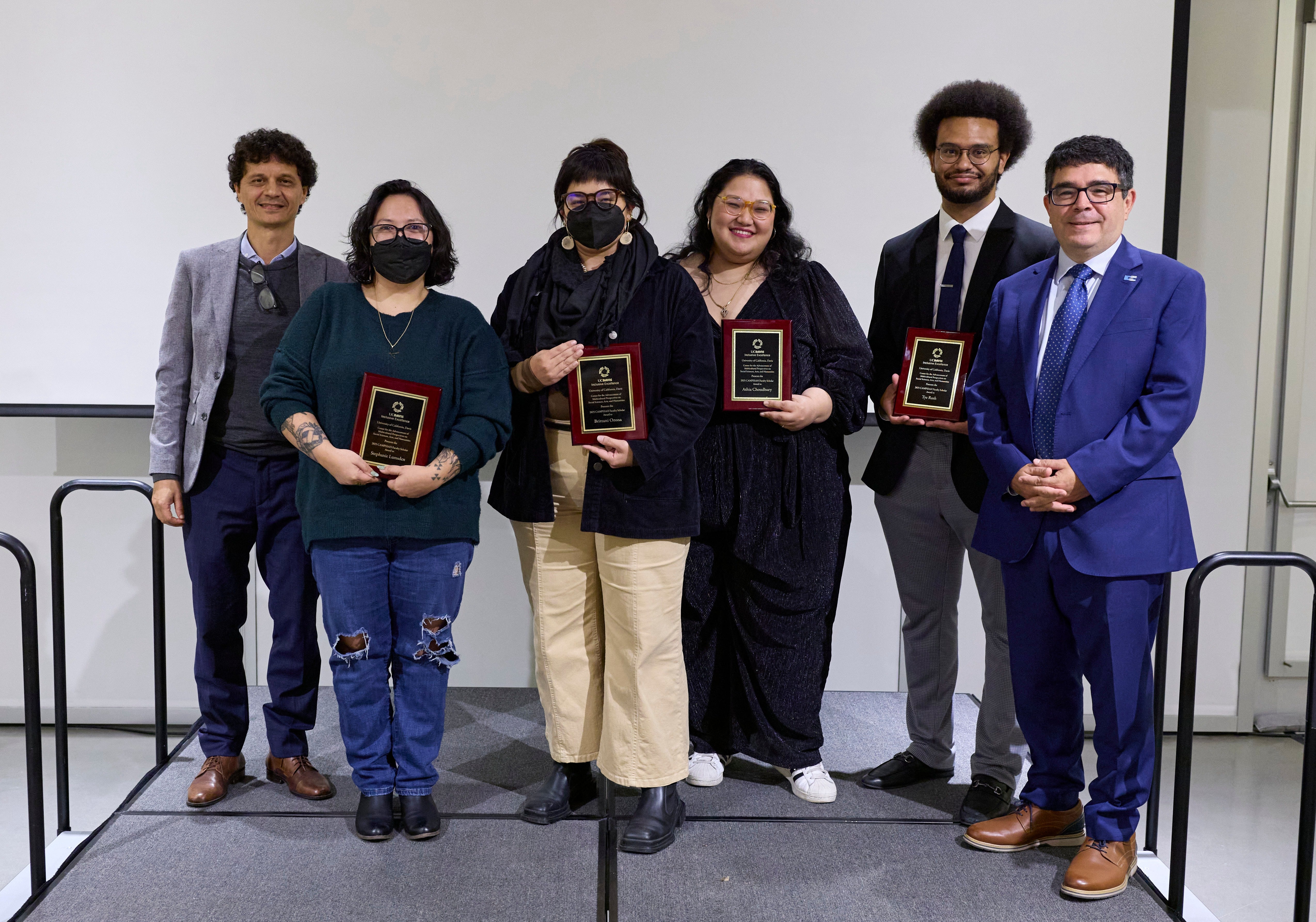 Six academics in professional dress stand in a row facing the camera on a stage in front of a blank projection screen. Several of the adults featured are holding plaques in their honor. 