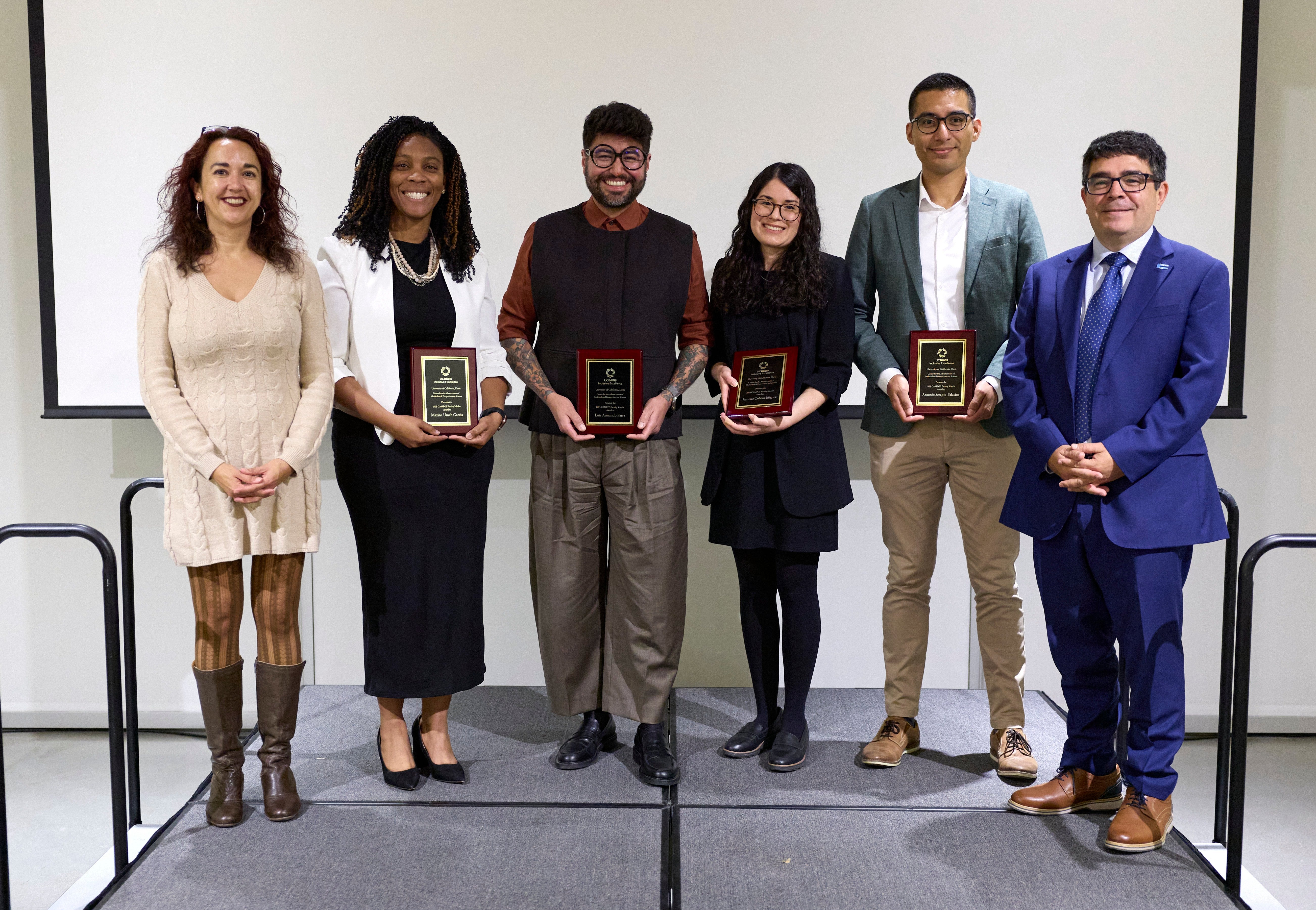 six scholars in professional dress stand atop a stage in front of a blank projection screen. several hold commemorative plaques, with all smiling and facing the camera. 