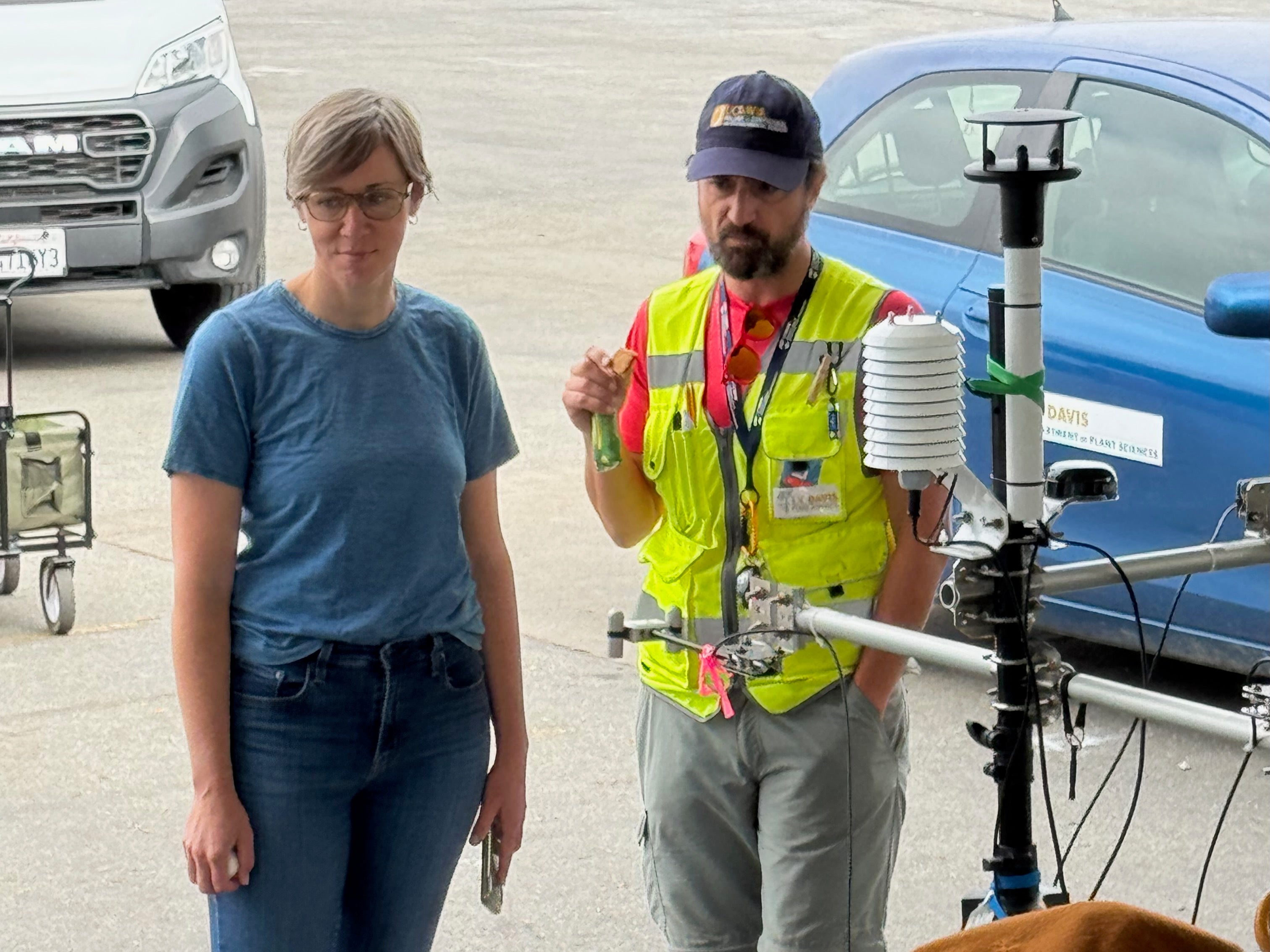 Kirsten Schwarz in blue short-sleeved shirt and jeans stands with Alessandro Ossola in yellow jacket and UC Davis hat with blue car and blacktop in background