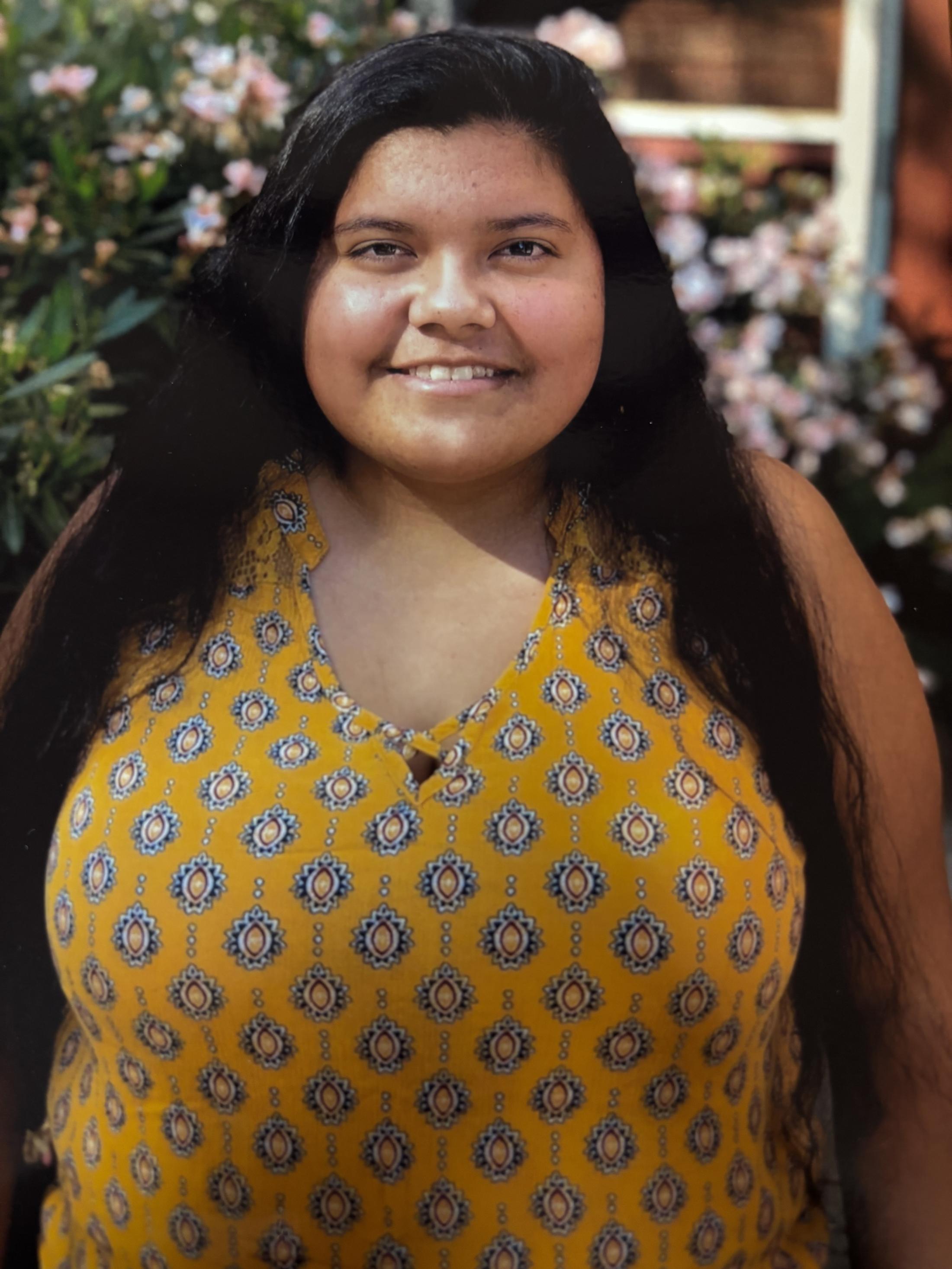 Jazmin Chavez, a UC Davis student, stands smiling outdoors in front of blooming flowers. She wears a sleeveless yellow patterned top, and her long black hair falls over her shoulders. The background is softly blurred with greenery and a building window visible.