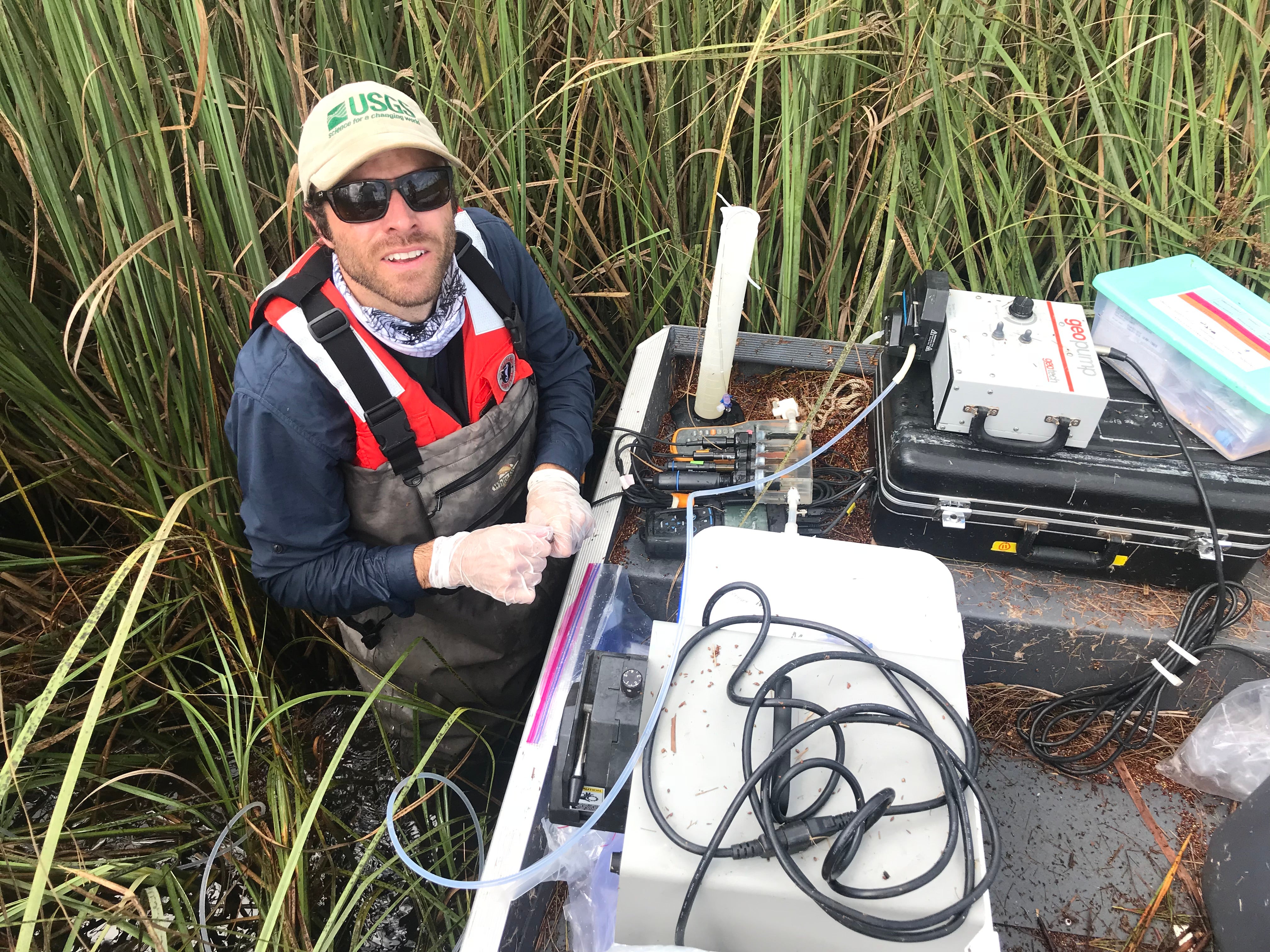Brett Poulin in ballcap, waders, clear gloves and sunglasses with scientific equipment among the water and plants of Florida Everglades