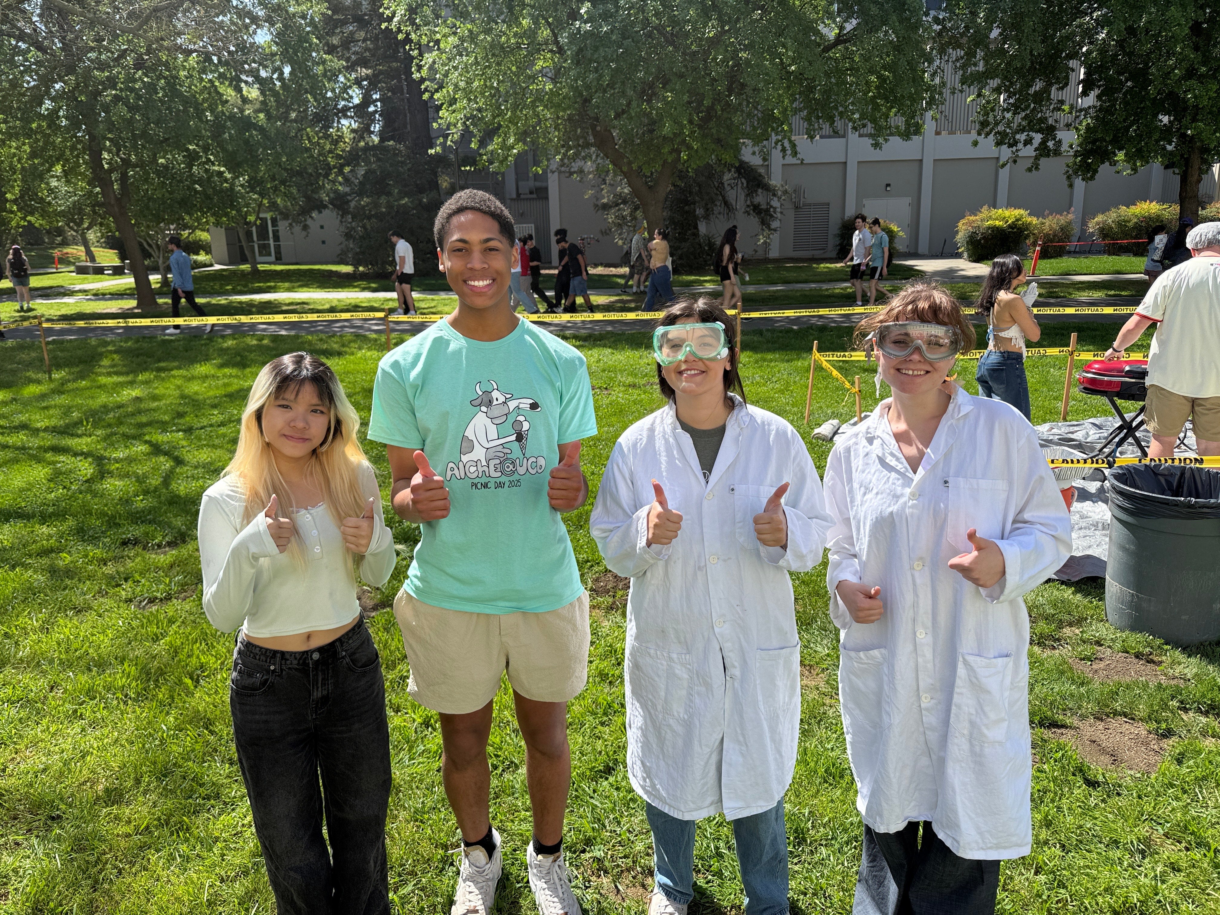 In an outdoor lawn setting, four students stand in a line smiling and giving thumbs up signs to the camera.