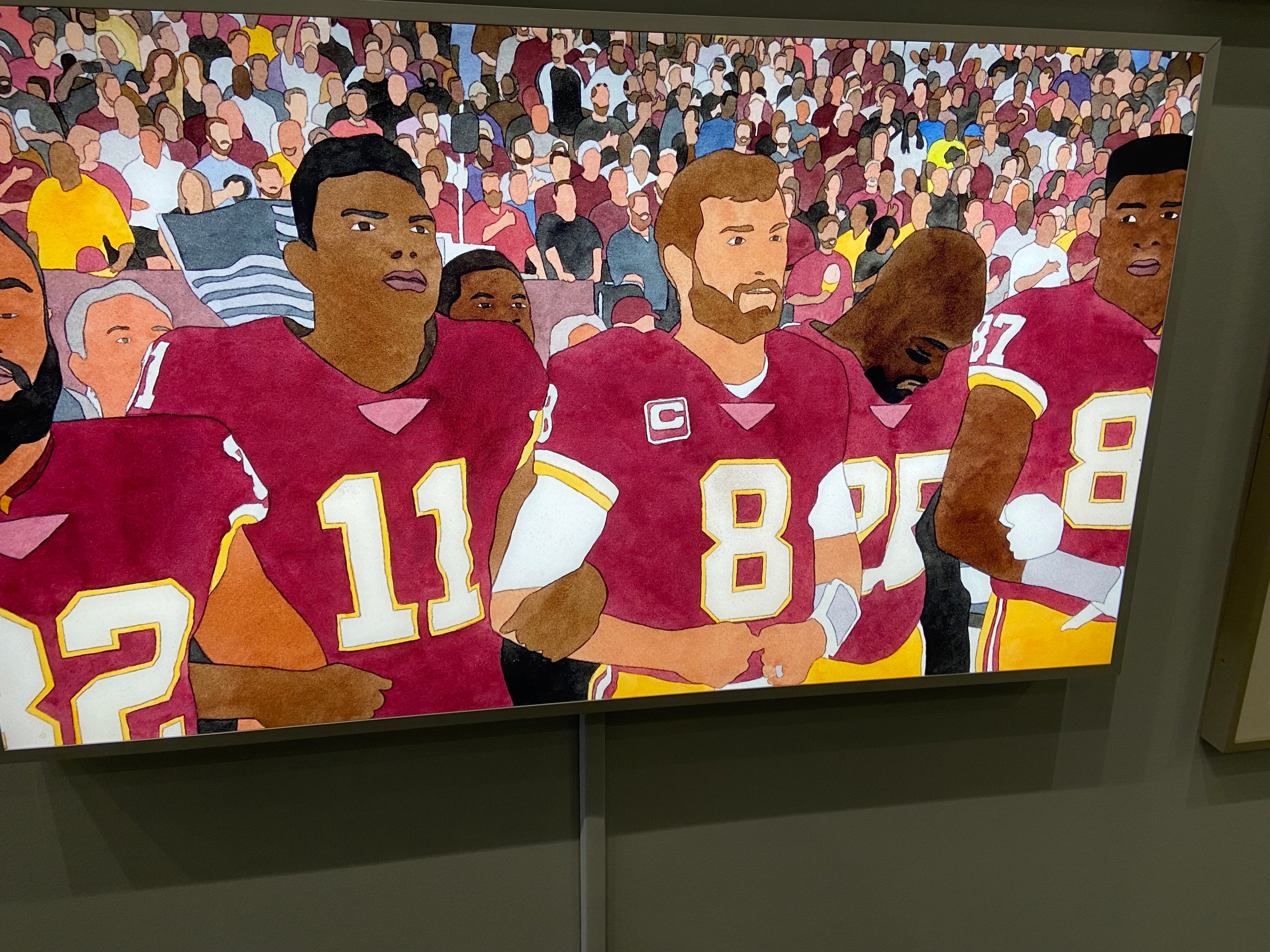 Group of football players in maroon uniforms standing together, with a cheering crowd in the background.