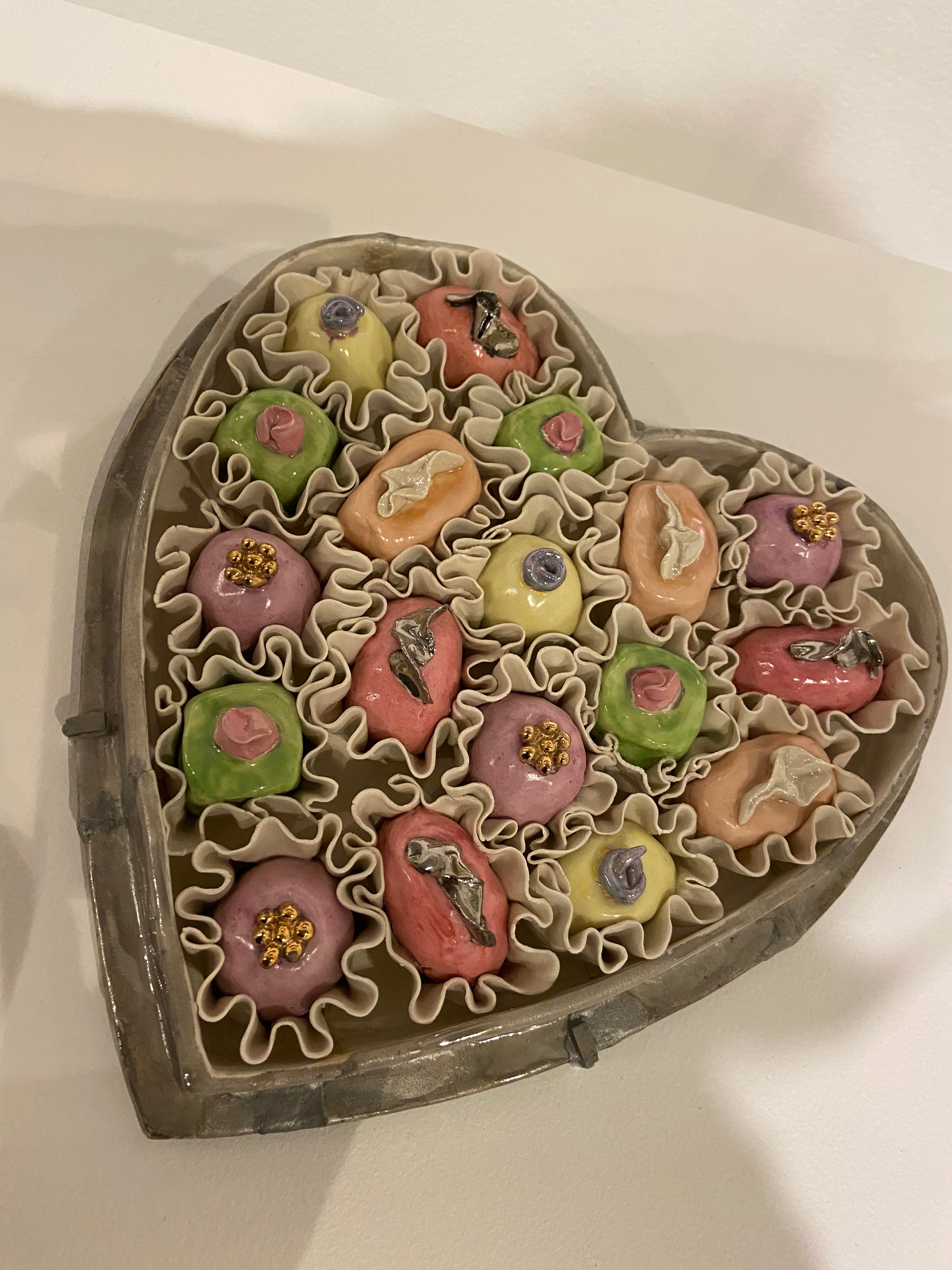 Ceramic depiction of heart-shaped box filled with colorful, decorative floral-shaped confections.