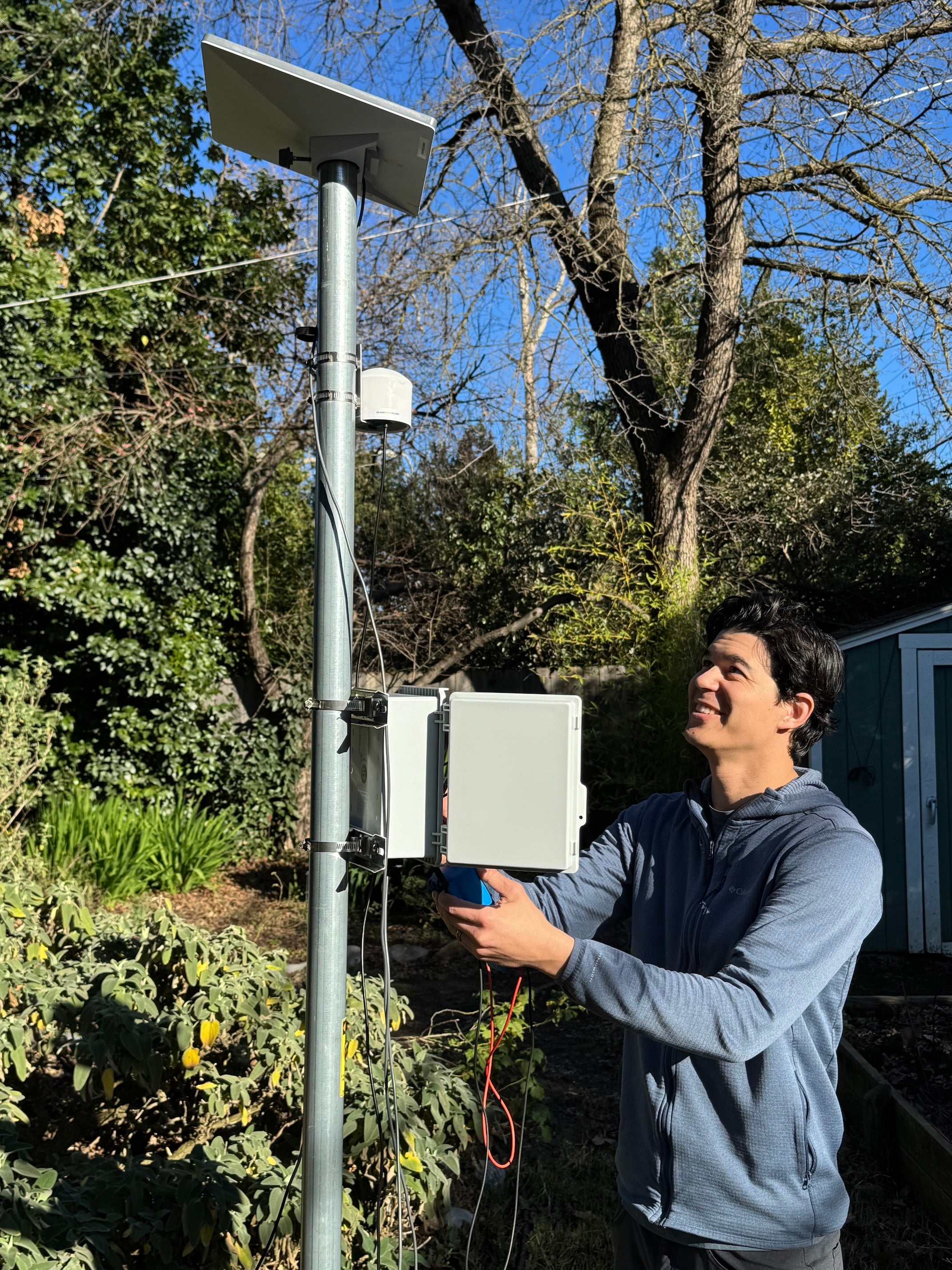 Christopher Wallis with dark hair looks up at air monitor installed on pole with solar panels. Trees and blue sky in background.