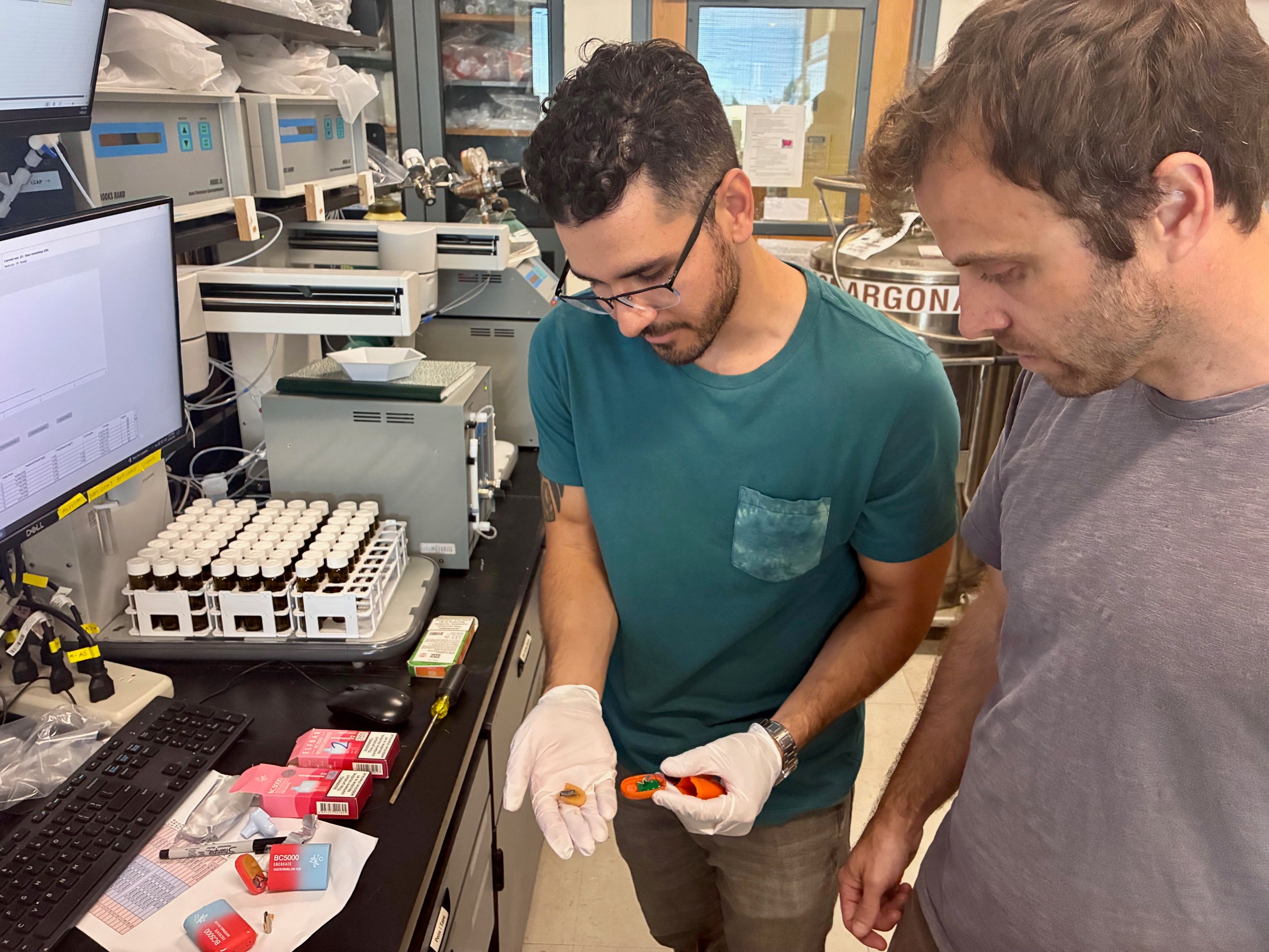 Mark Salazar in lab holds a vaping pod near a counter showing other vaping pods and scientific gear as Brett Poulin looks down at it
