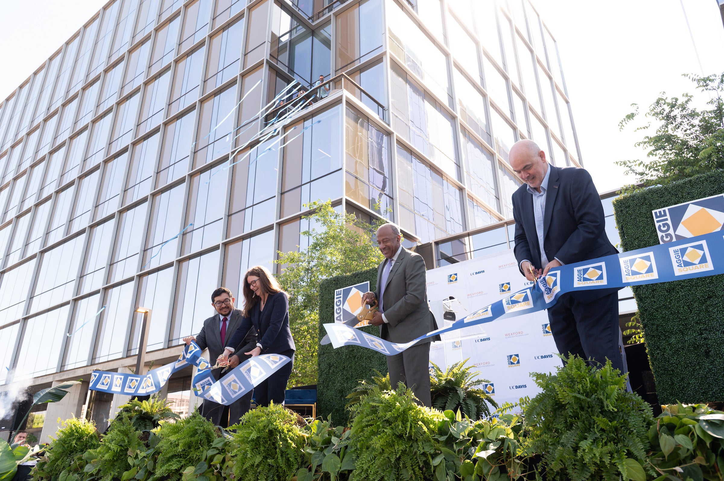 Outdoor setting, large glass building in background. Four people stand in a row cutting a ribbon into pieces. 