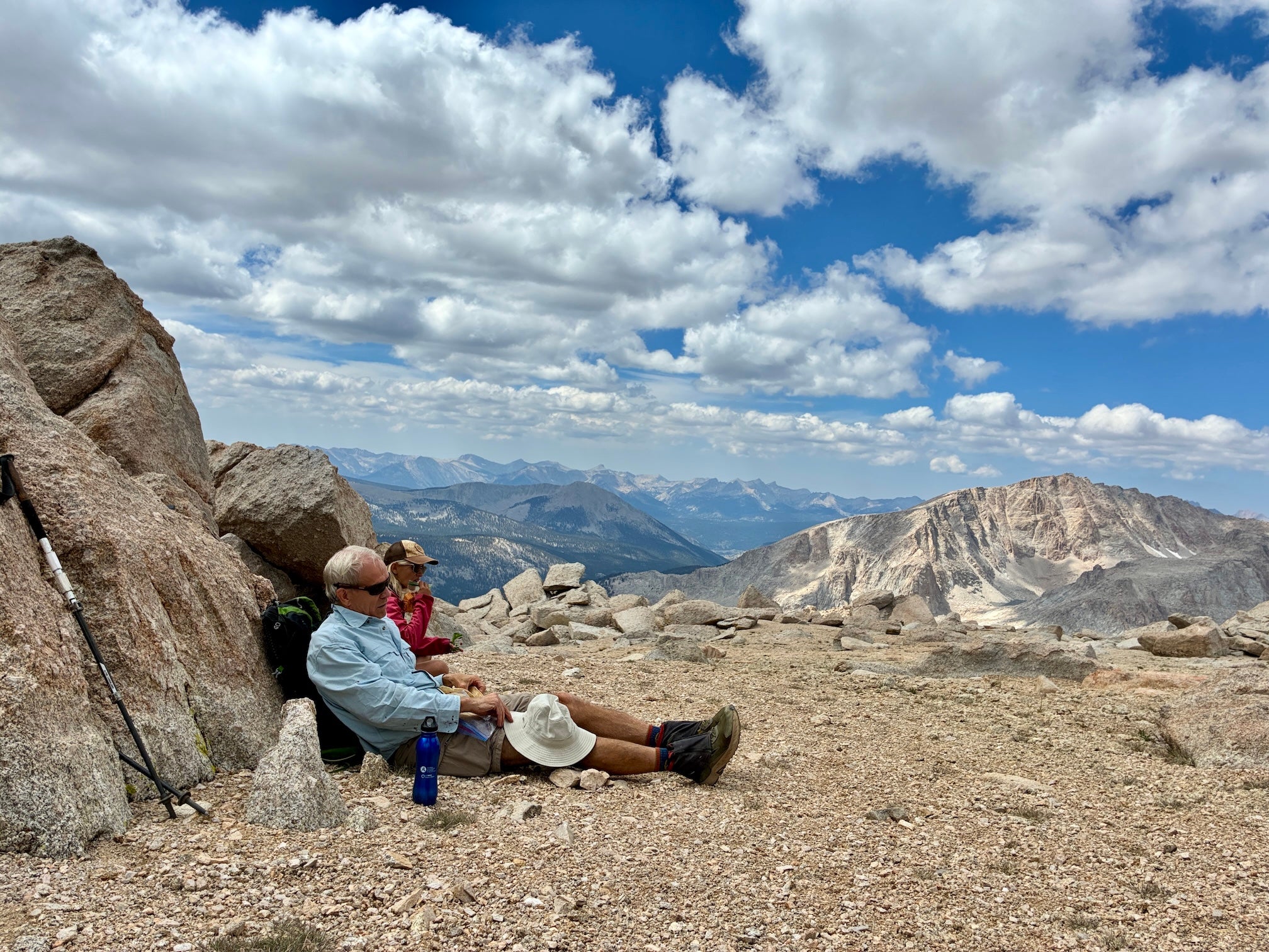 Man in blue shirt and woman in red shirt slump resting against boulder with vista or Sierra mountains in background