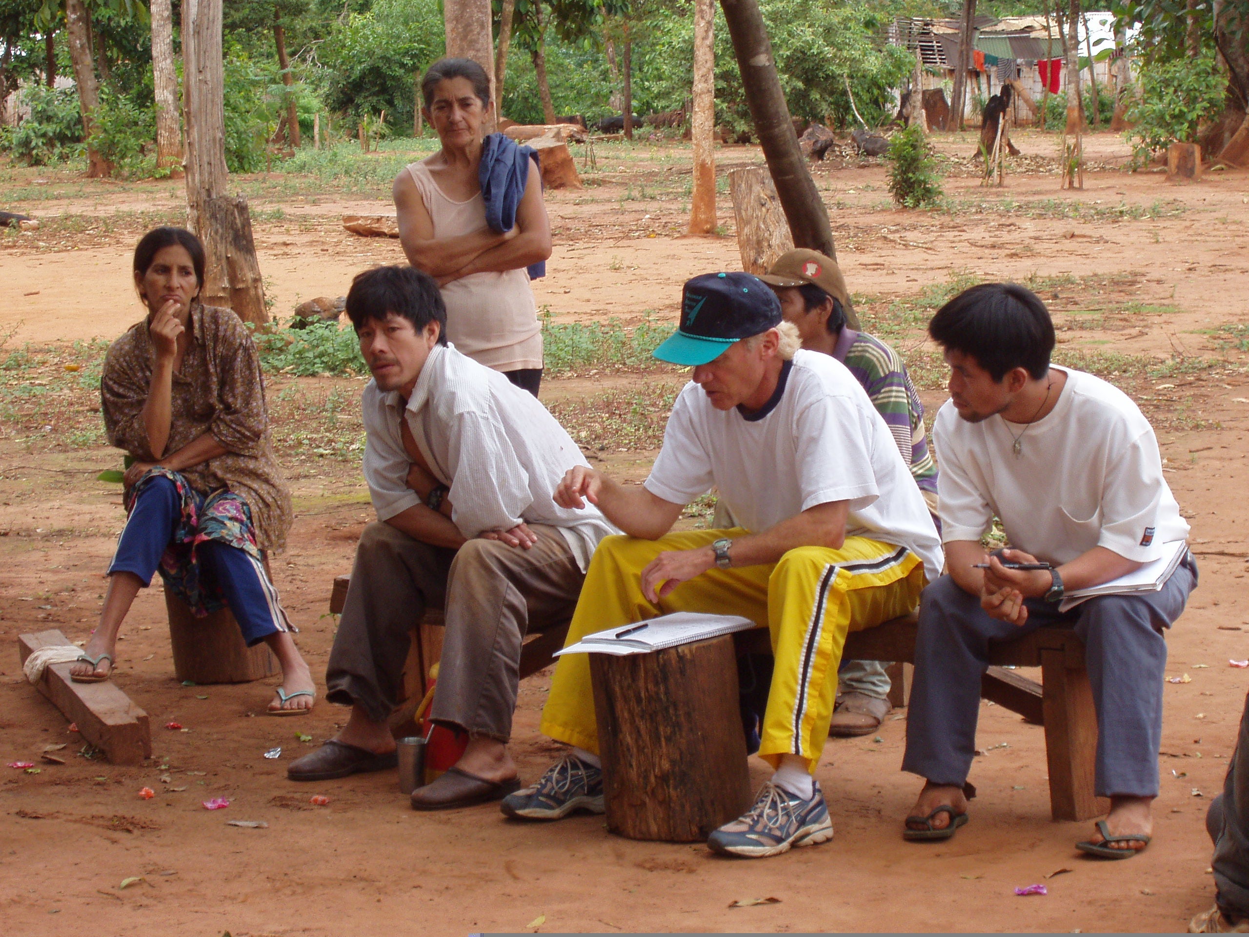 Four people seated outdoors. Second from right is a European male wearing yellow trackpants, blue ball cap and white t-shirt. On a tree stump in front of him is a clipboard and some papers. 