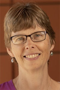 Smiling woman with short brown hair and glasses, wearing earrings and a purple top.