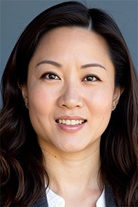 A woman with long dark hair smiles warmly, wearing a gray blazer against a neutral background.
