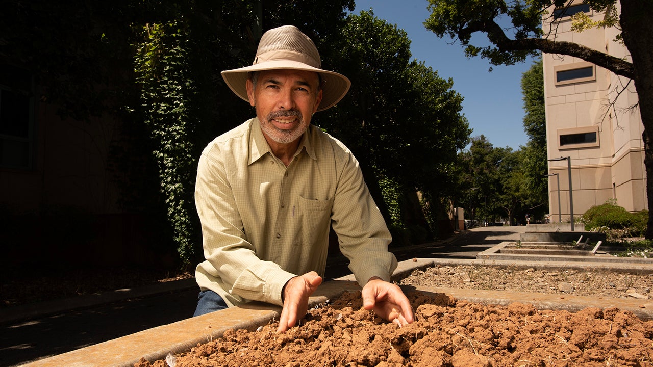 Kneeling beside a raised planting bed, Jorge Mazza Rodrigues gathers some dirt in his hands.