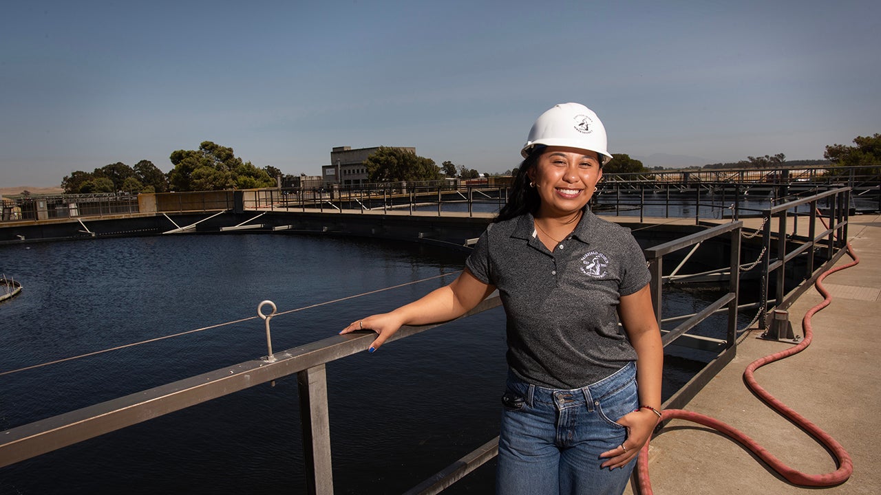 Wearing a hardhat, Krystle Catamura stands with large reservoirs behind her