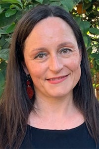 A woman with long dark hair smiles warmly, surrounded by green foliage.