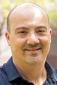 Smiling man with a bald head wearing a dark shirt, outdoors with blurred greenery in the background.