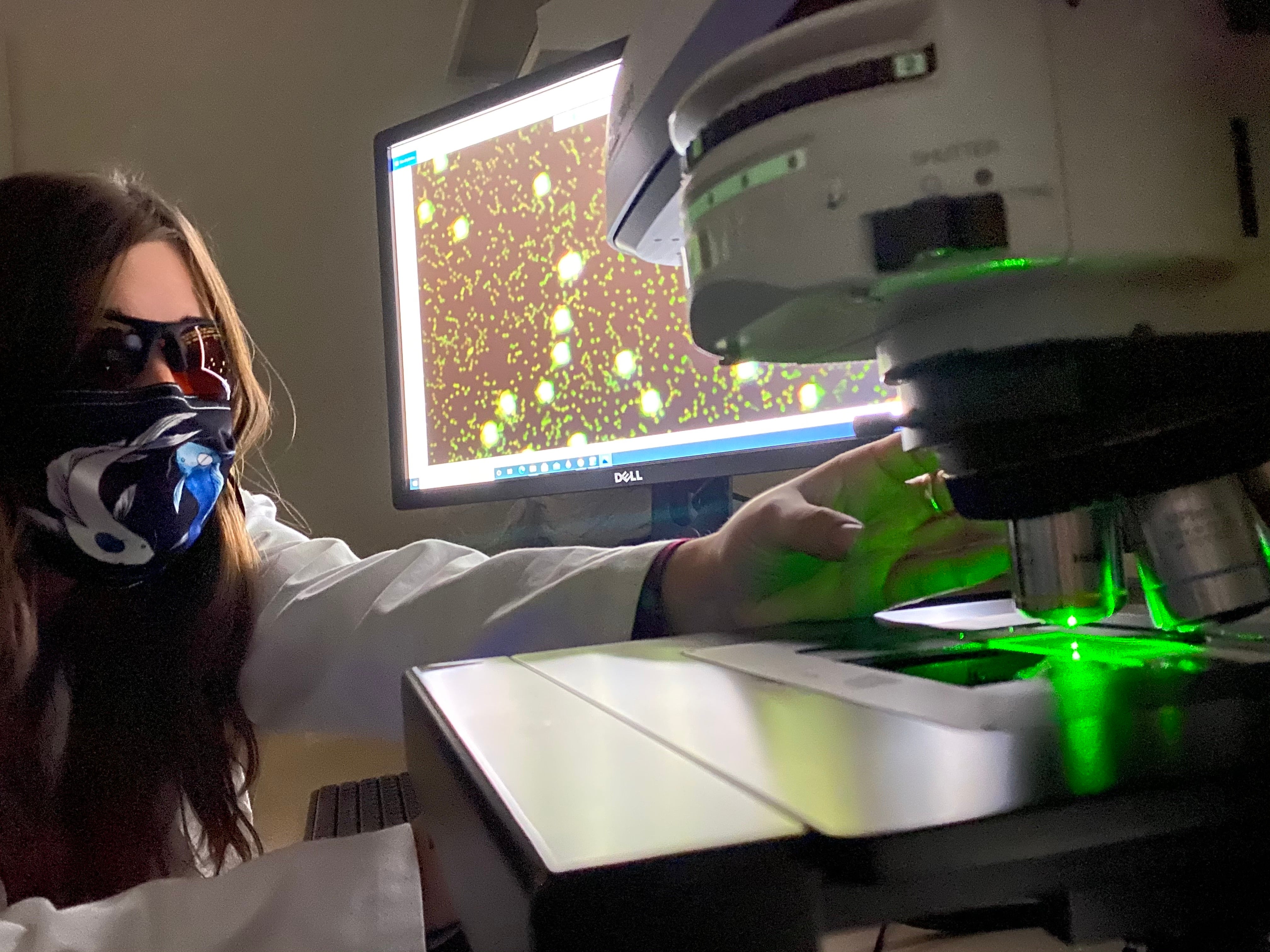 A woman scientist in mask at computer and scientific instrument with green laser