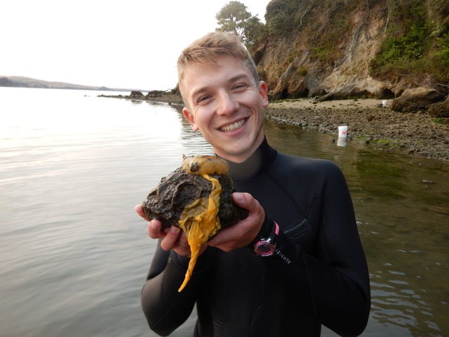 White male scientist in wet suit holds sea lemon and smiles at camera with ocean bay in background