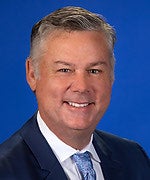 Headshot of a man wearing a suit and tie and smiling at the camera, against a blue background