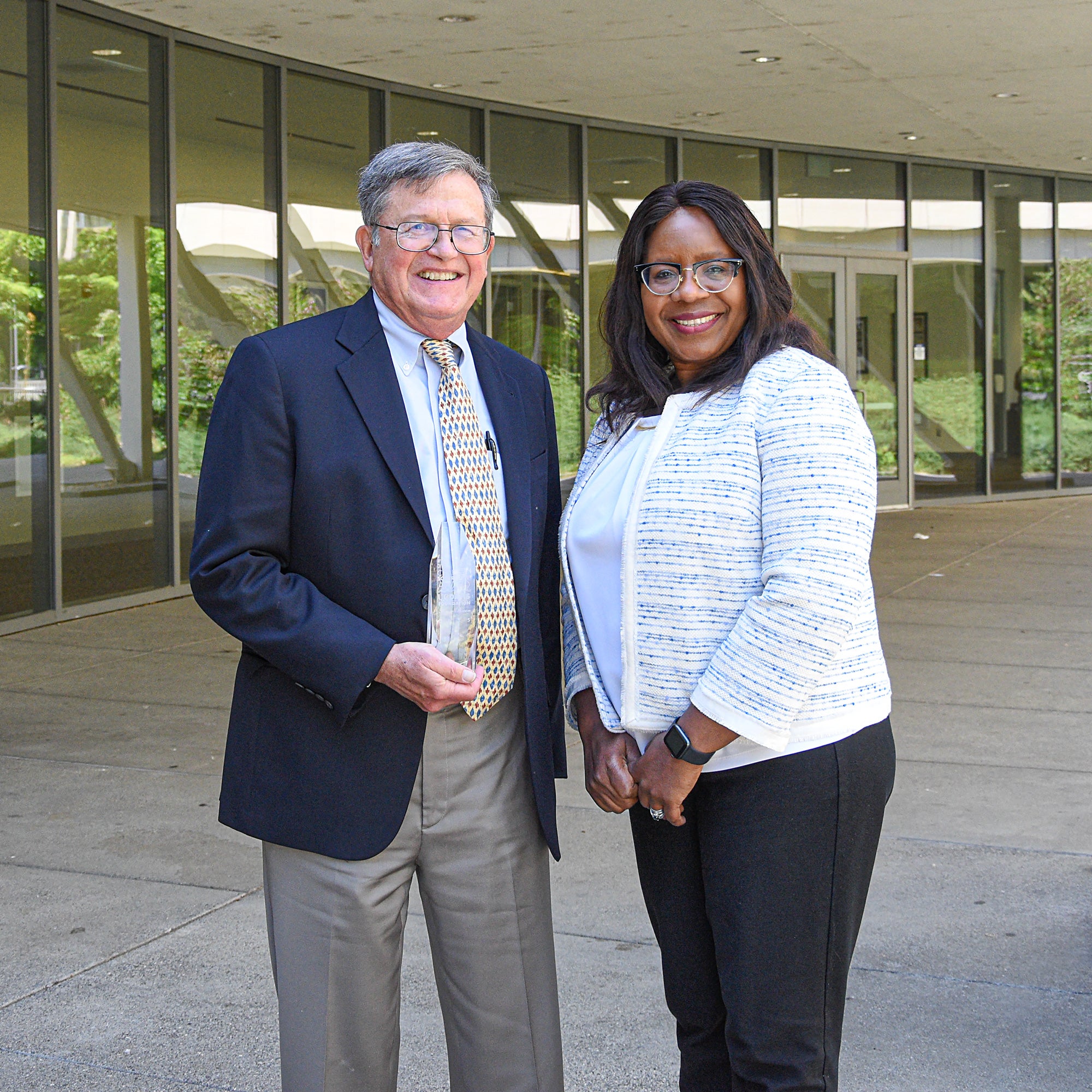 A man and a woman stand outside a venue in professional wardrobes, smiling and standing next to each other