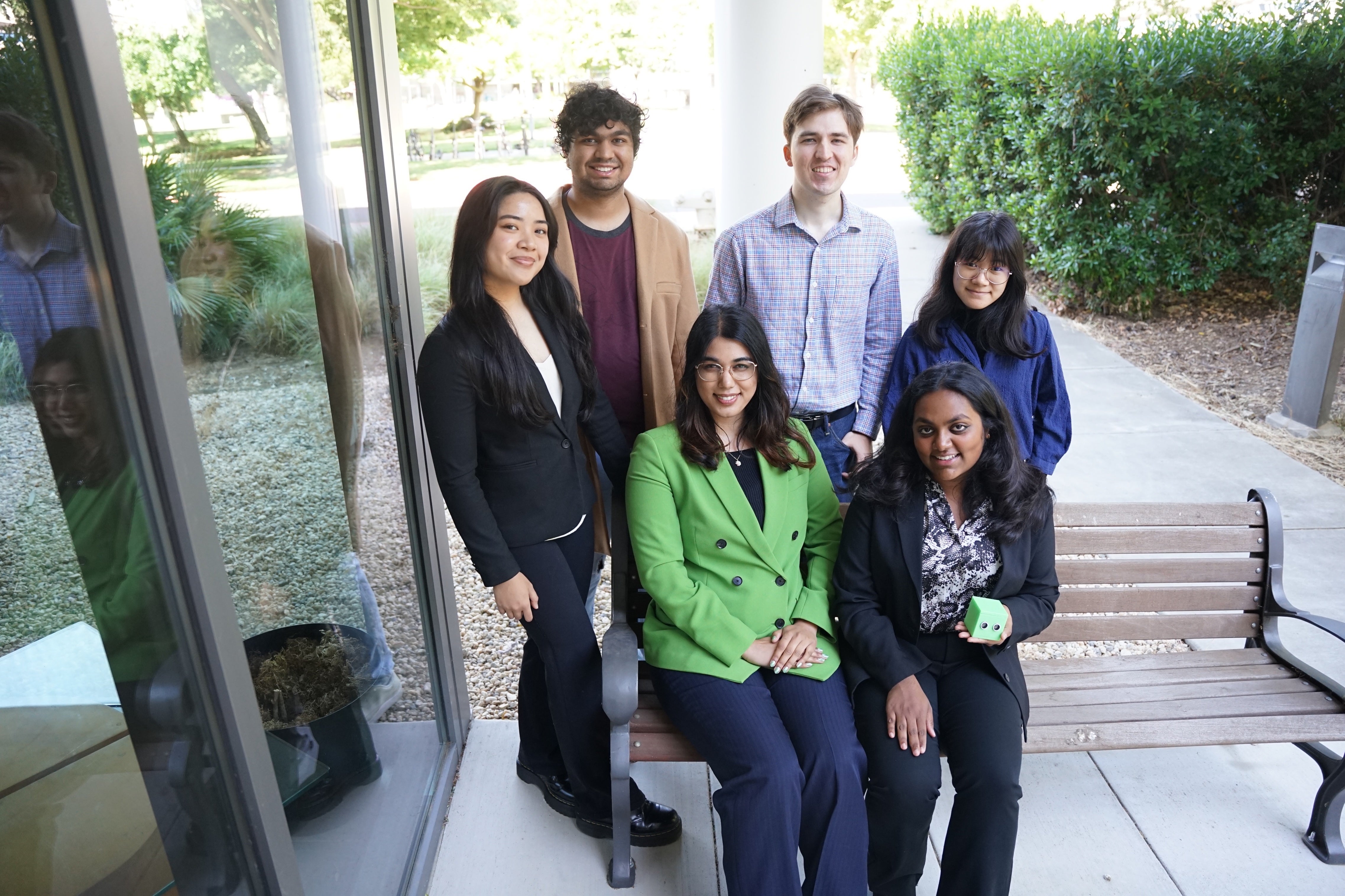 Six students are posed on and behind a bench, with one holding a green cube with two round "eyes" in the front